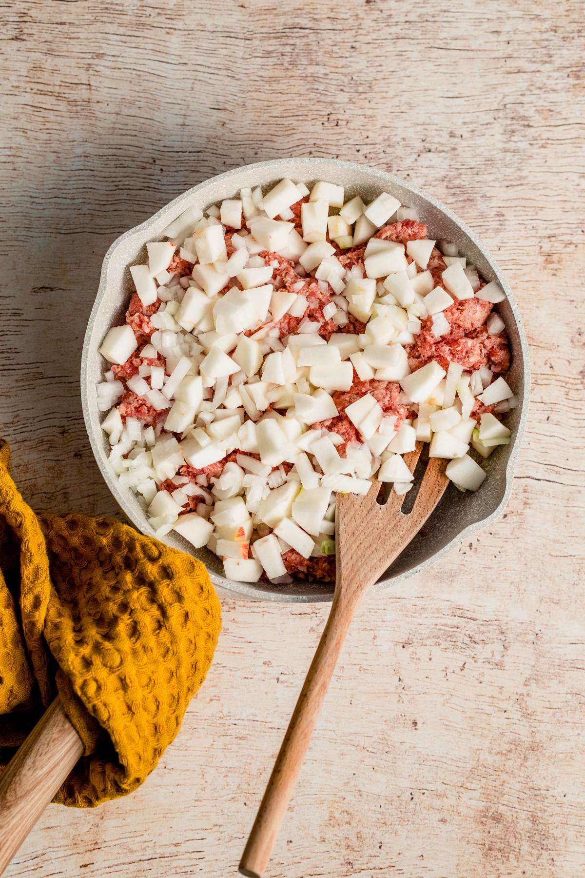 A pan of chopped onions and ground meat with a wooden spatula sits next to a yellow textured cloth, perfect as the base for a breakfast casserole with apples—an inviting choice for Thanksgiving or Christmas breakfast.