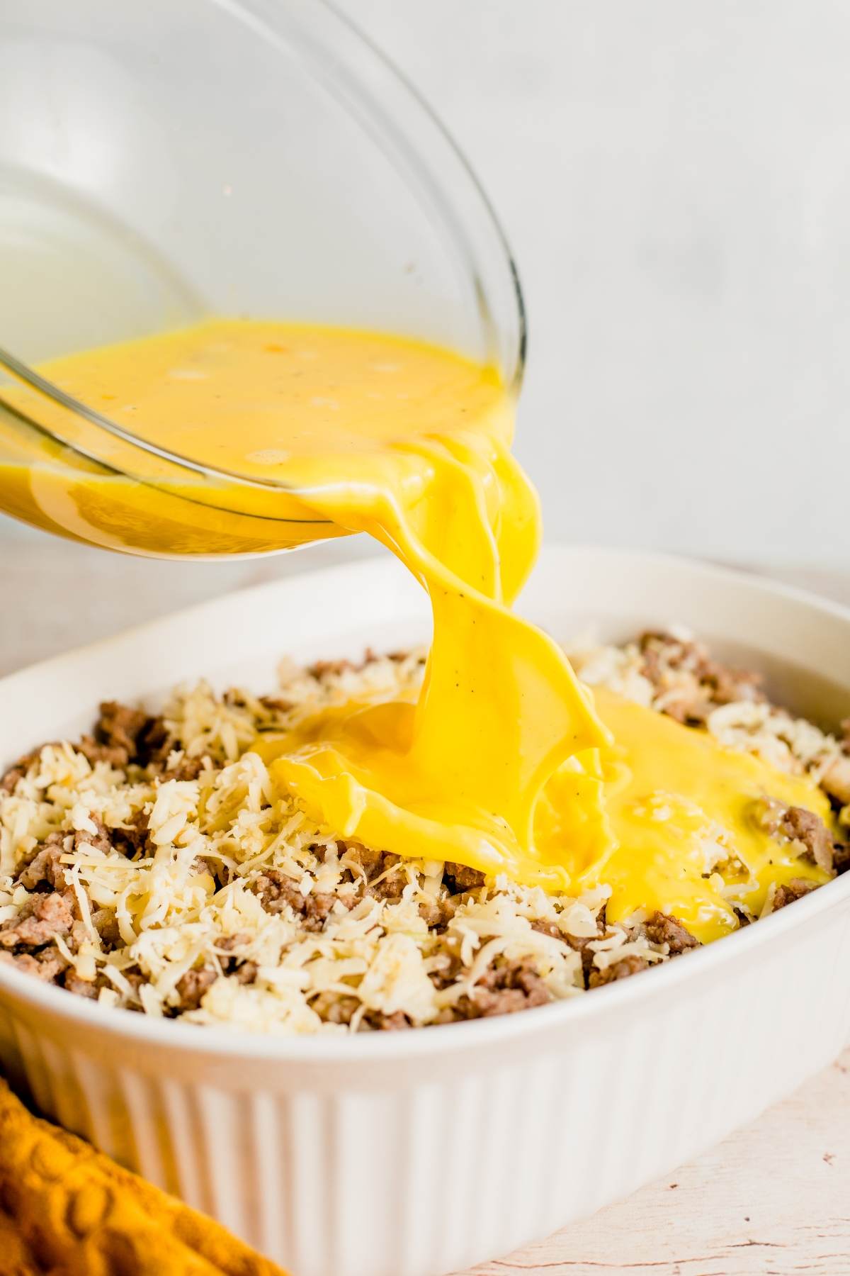 Beaten eggs are poured over shredded cheese and ground meat in a white baking dish, creating the perfect Thanksgiving or Christmas breakfast casserole.