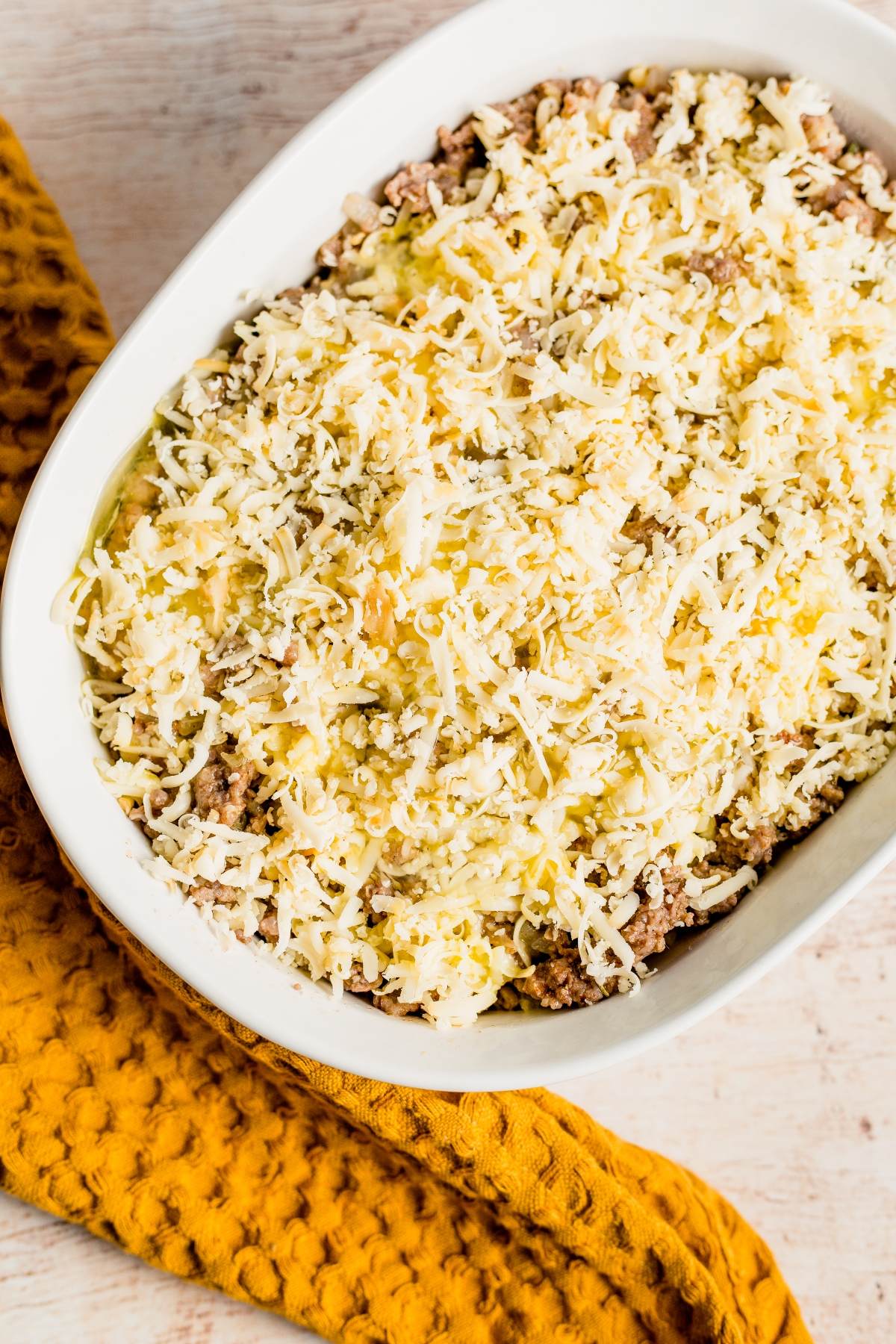 A casserole dish filled with shredded cheese over a savory meat mixture sits beside a textured yellow cloth—a perfect easy family brunch recipe or Thanksgiving or Christmas breakfast.