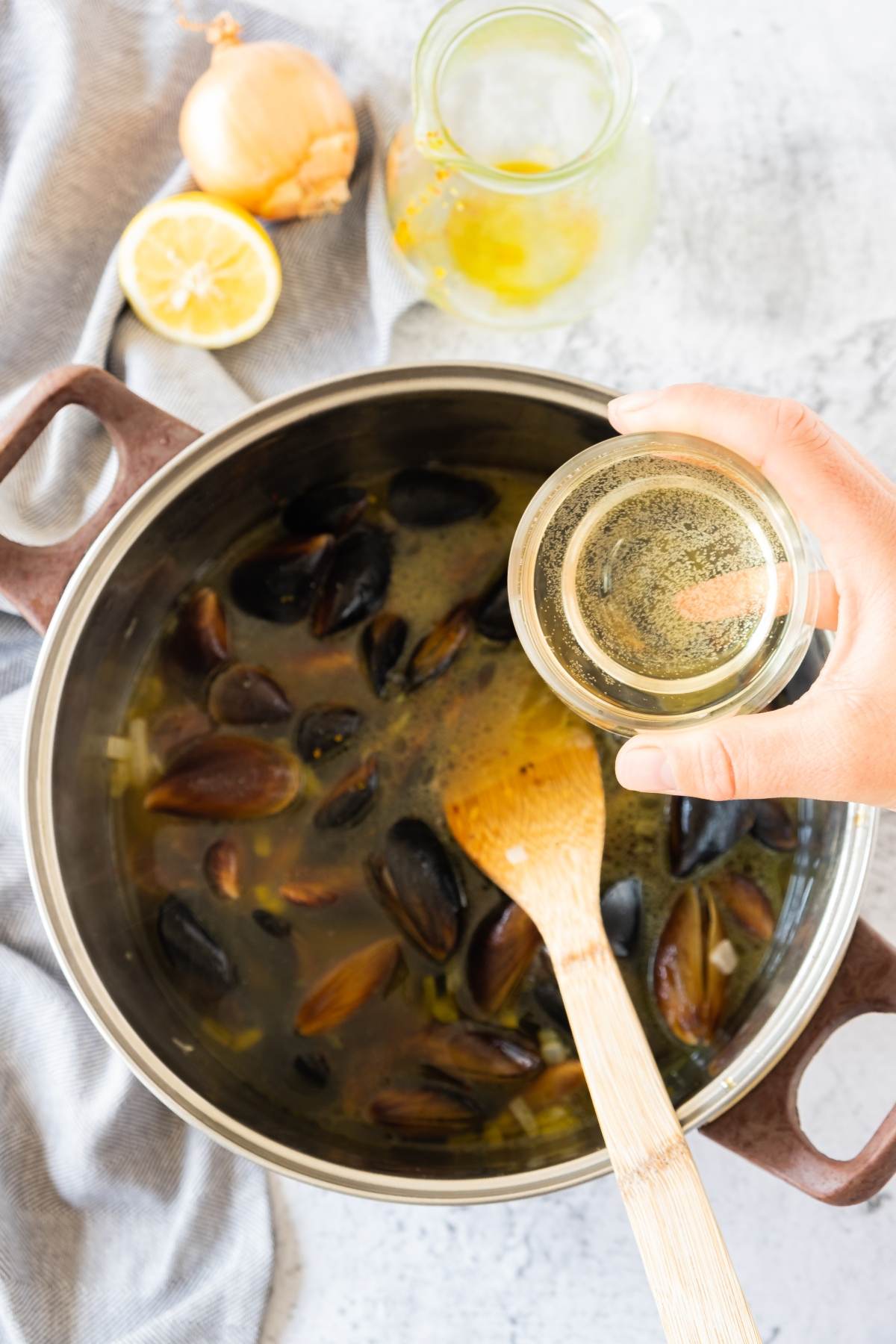 A hand pours white wine into a pot of mussels and broth with a wooden spoon, onion, and lemon nearby—capturing the essence of an easy seafood paella recipe.
