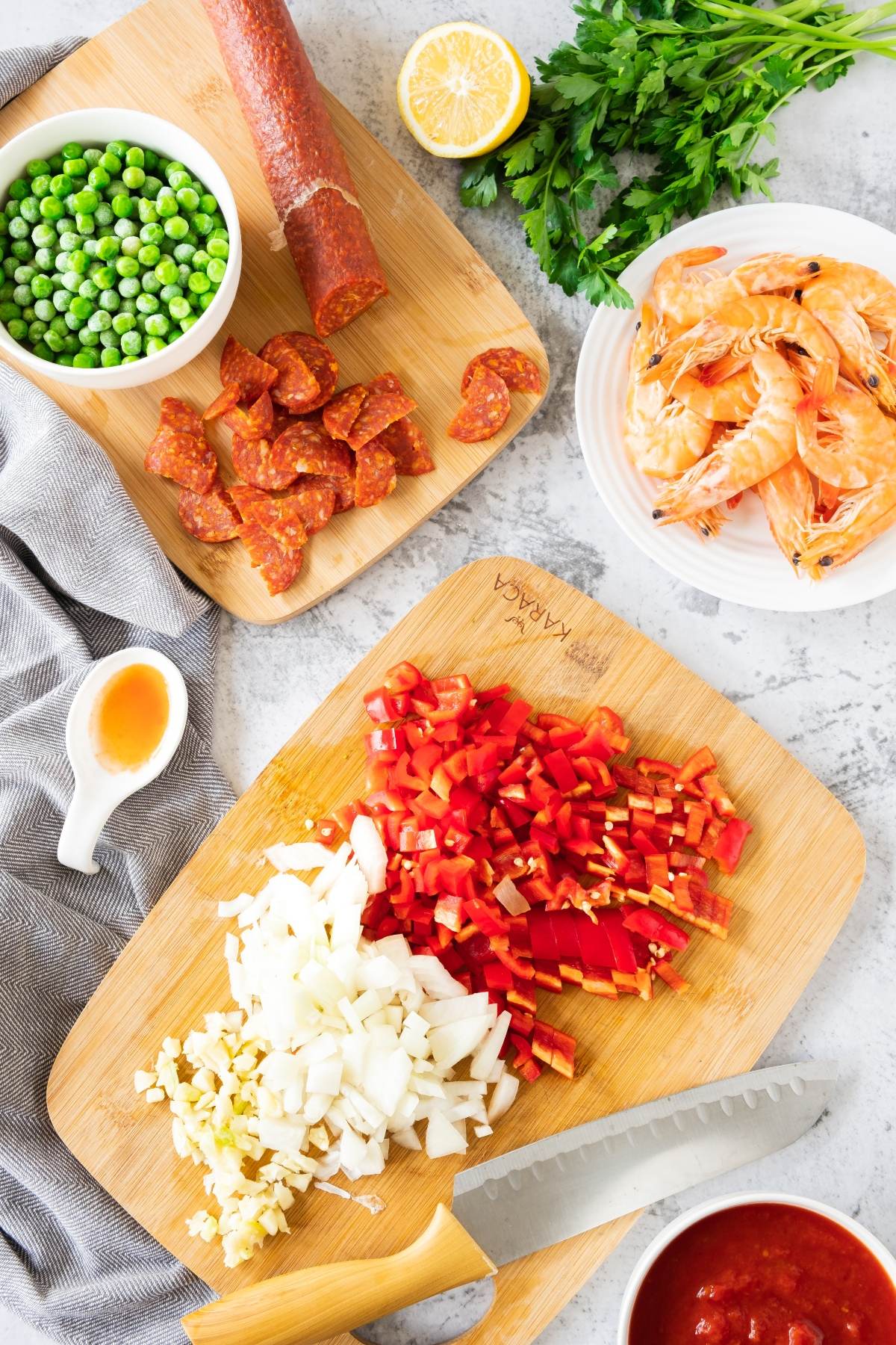 Chopped onions, garlic, and red peppers on a board with shrimp, peas, chorizo, herbs, lemon, and sauce nearby—perfect ingredients for traditional Spanish paella or a flavorful Seafood Chorizo Paella.