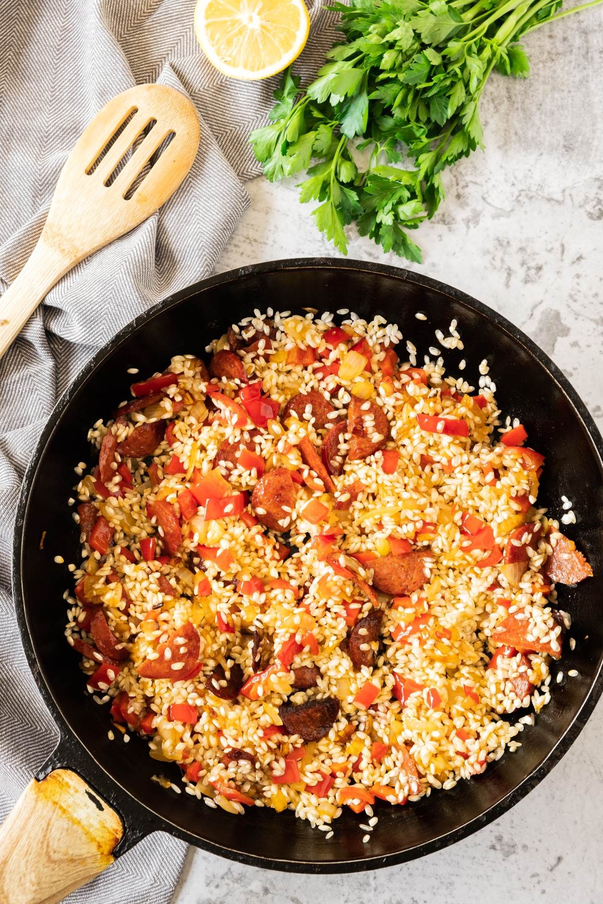 A skillet with rice, sliced sausage, red peppers, and herbs—this dish captures the vibrant flavors of seafood and chorizo paella—served next to parsley, lemon, and a wooden spoon.