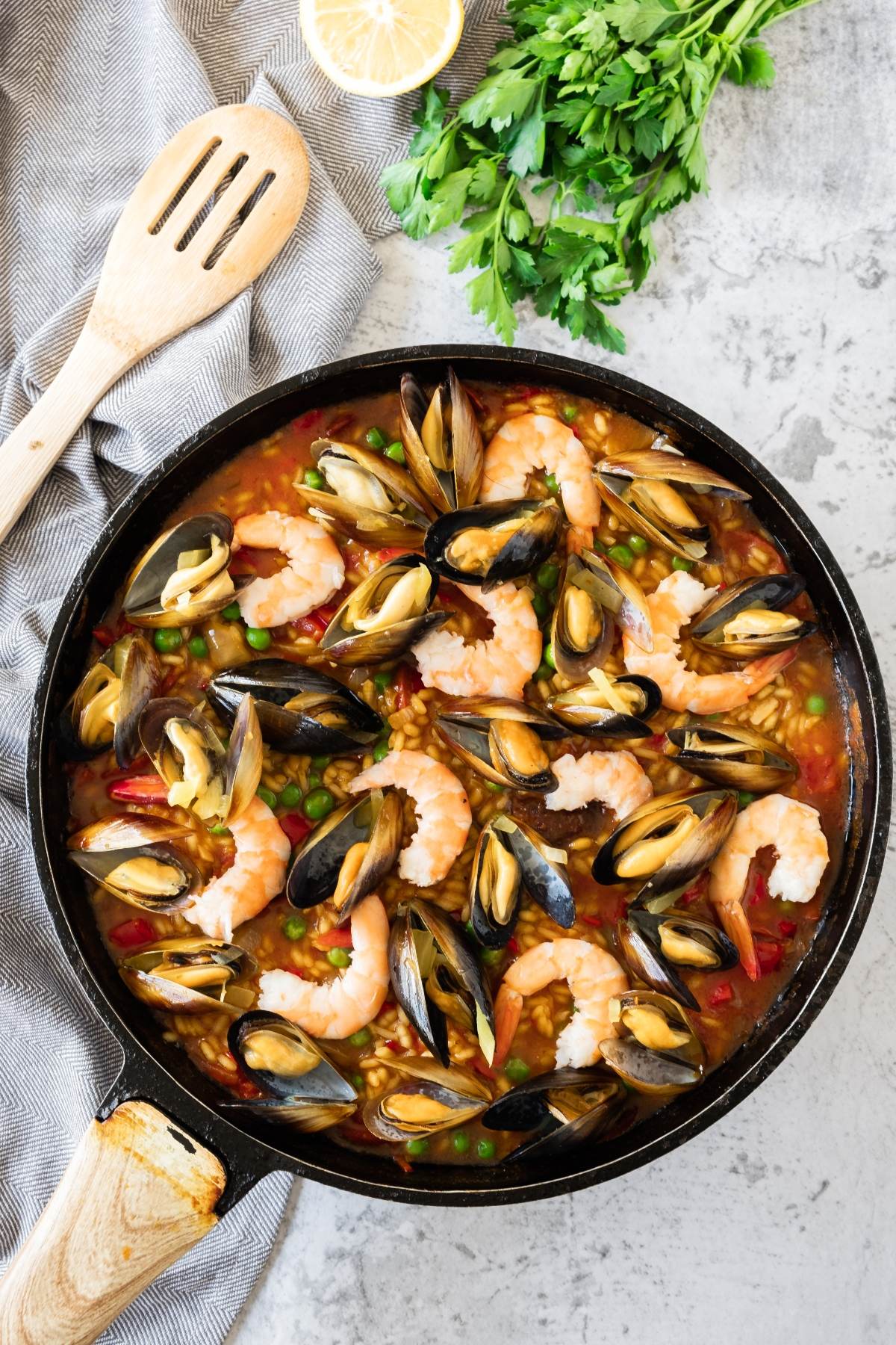 A pan of traditional Spanish paella with shrimp, mussels, peas, and rice sits on a gray countertop beside herbs and a wooden spoon—an easy seafood paella recipe perfect for any occasion.