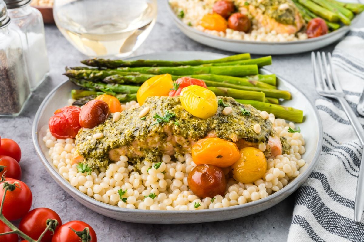 A plate of couscous topped with sheet pan salmon and asparagus, roasted tomatoes, and pesto, served with a glass of white wine.