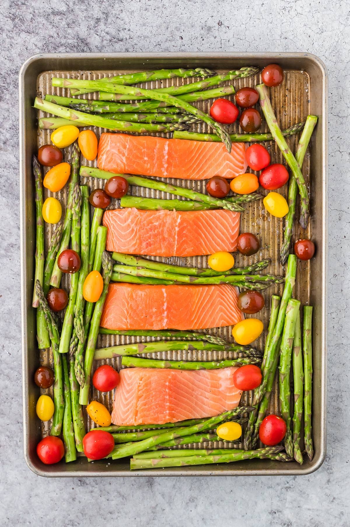 Four raw salmon fillets, asparagus stalks, and colorful cherry tomatoes on a baking sheet.