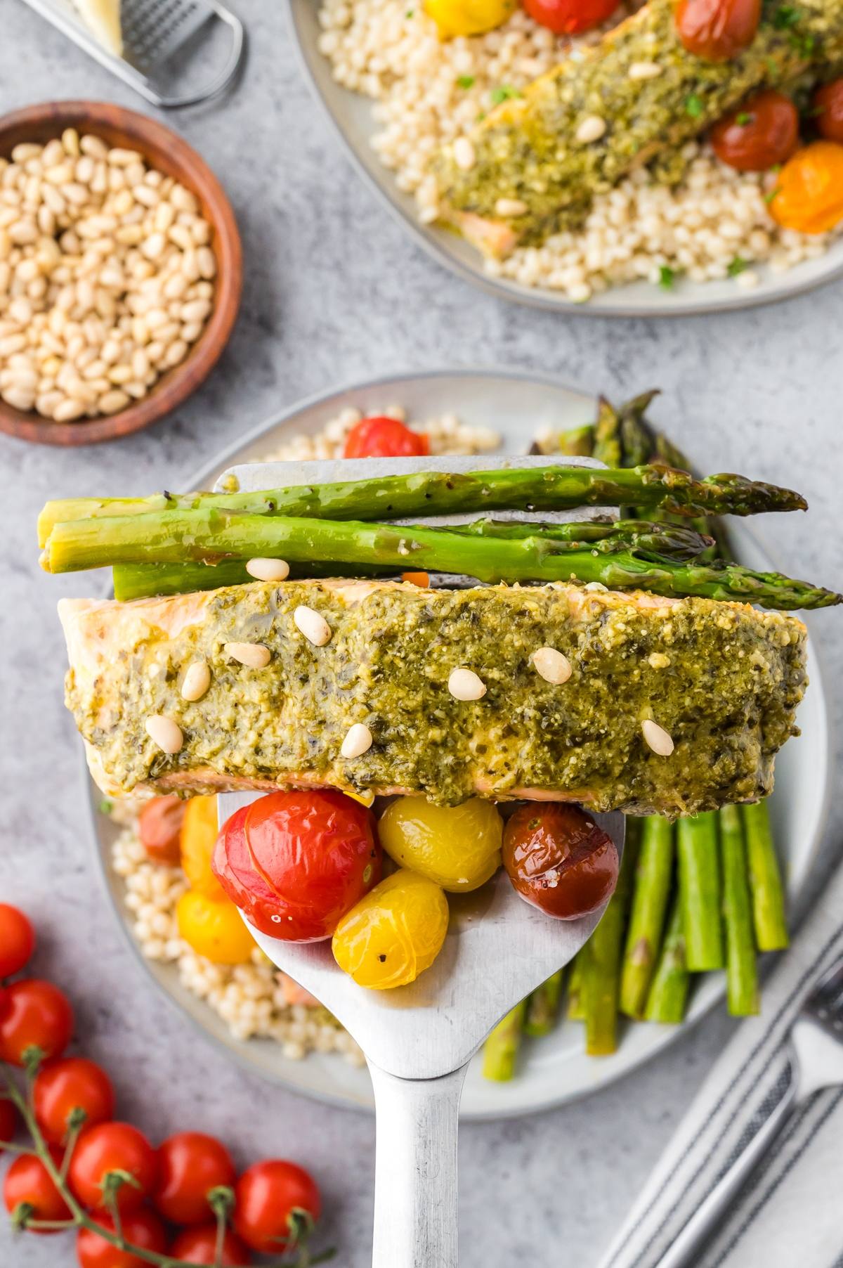 A spatula holds pesto salmon with asparagus and cherry tomatoes over a plate of couscous and vegetables.