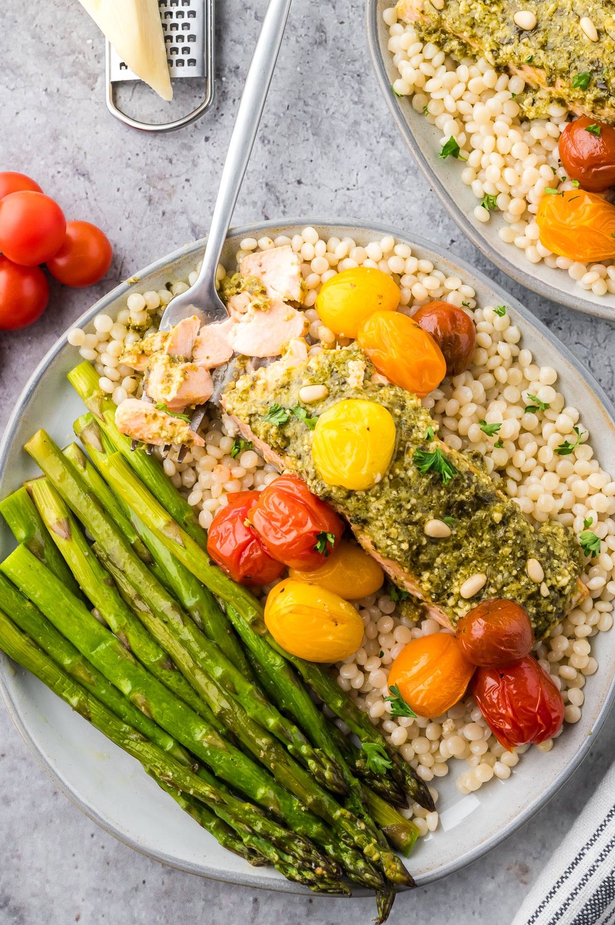 Plate of salmon with pesto, roasted tomatoes, asparagus, and pearl couscous on a gray surface.