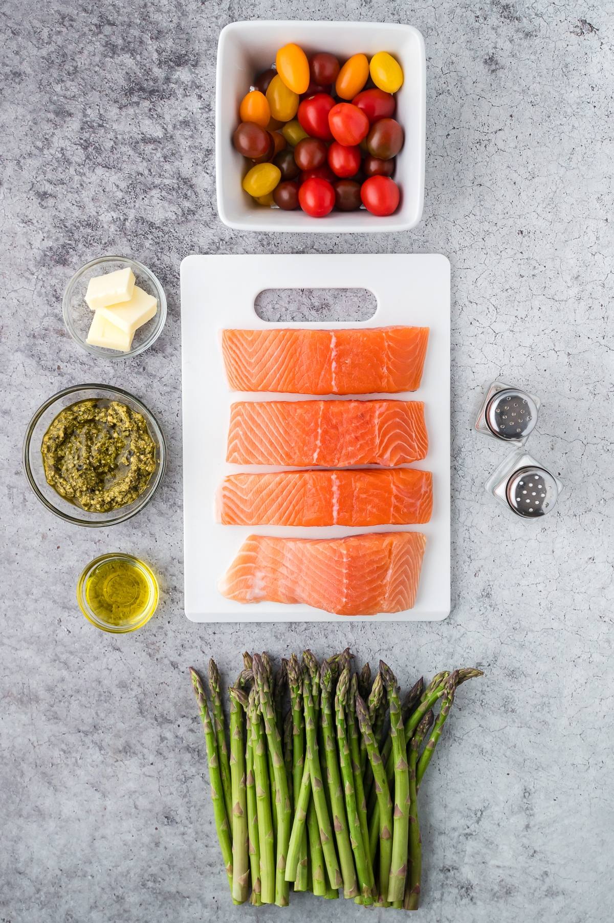 Four raw salmon fillets, asparagus, tomatoes, pesto, butter, olive oil, salt, and pepper on a gray surface.