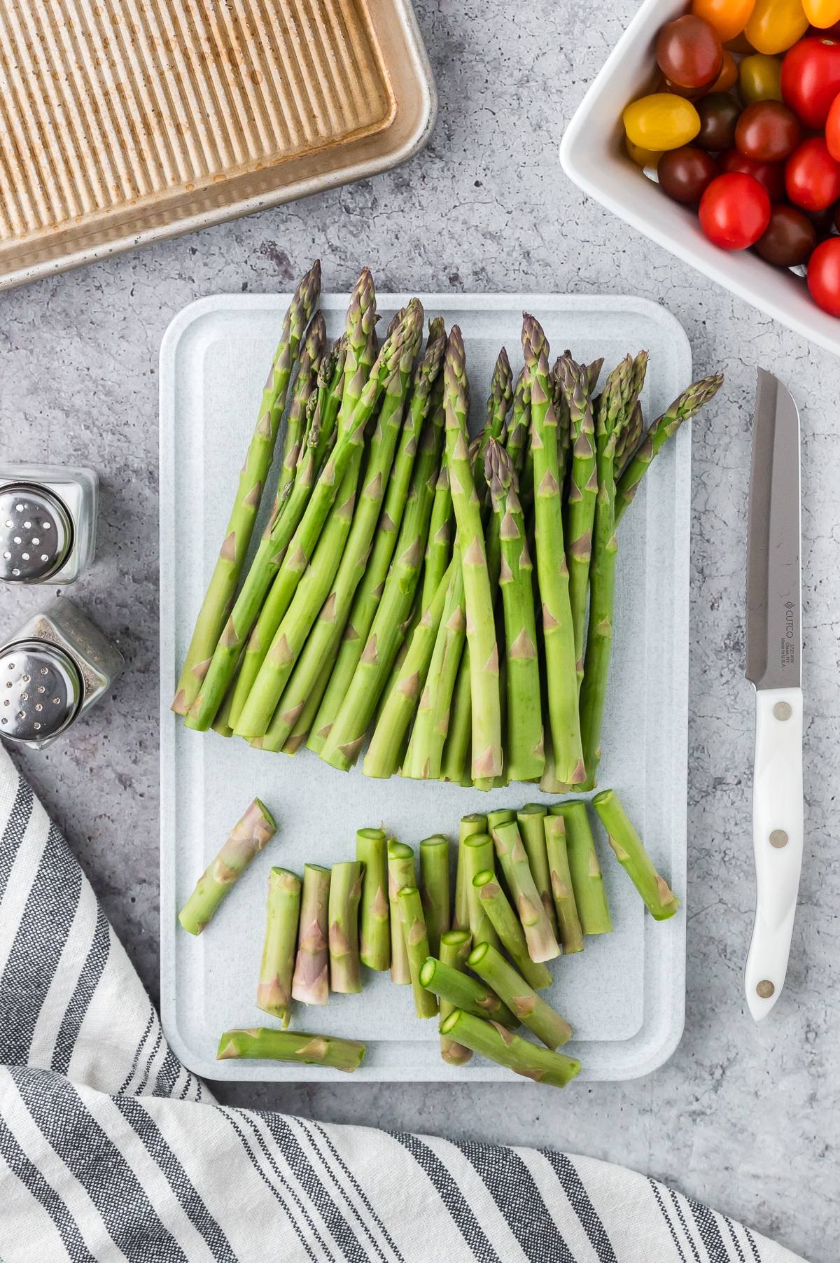Fresh asparagus spears and chopped ends on a cutting board, with a knife, salt, pepper, and cherry tomatoes nearby.