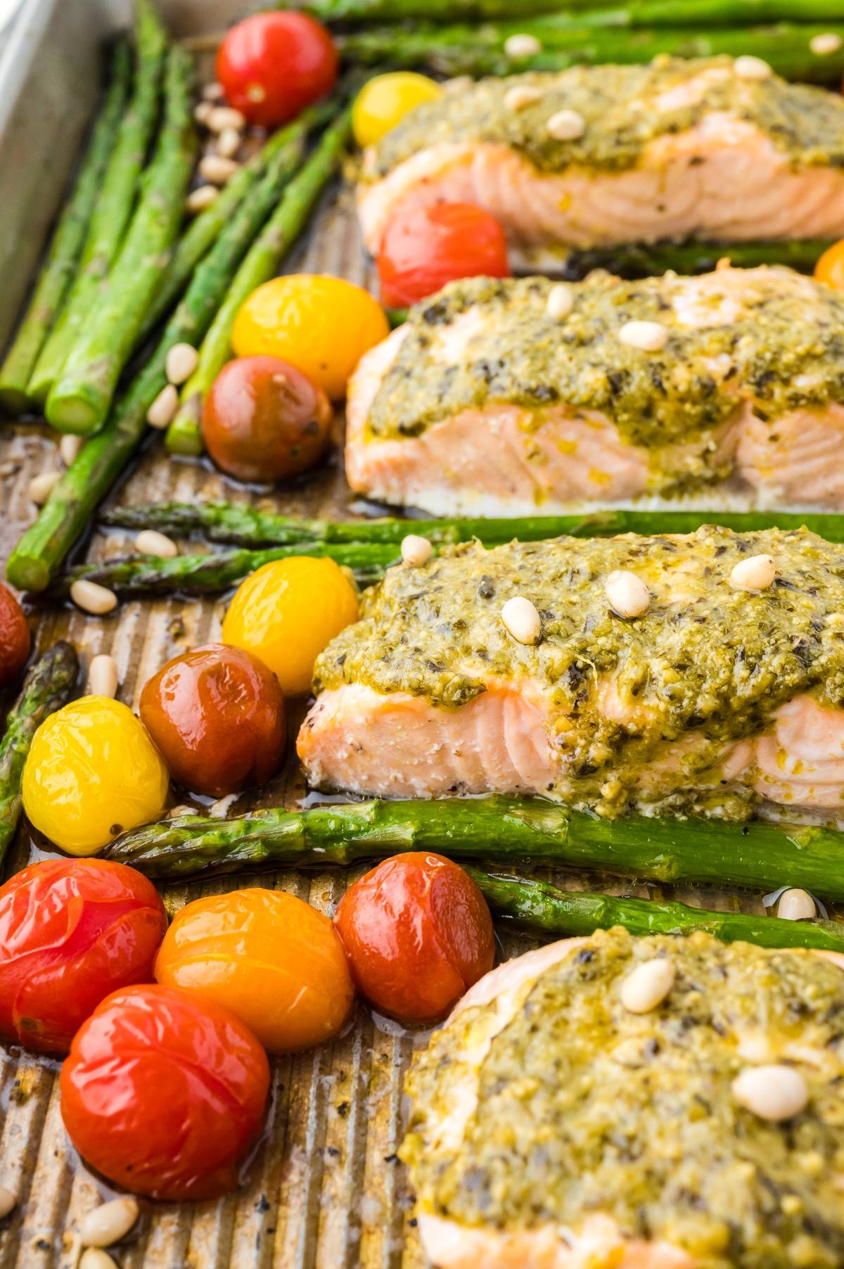 Baked salmon with pesto, asparagus, and roasted cherry tomatoes on a sheet pan, topped with pine nuts.