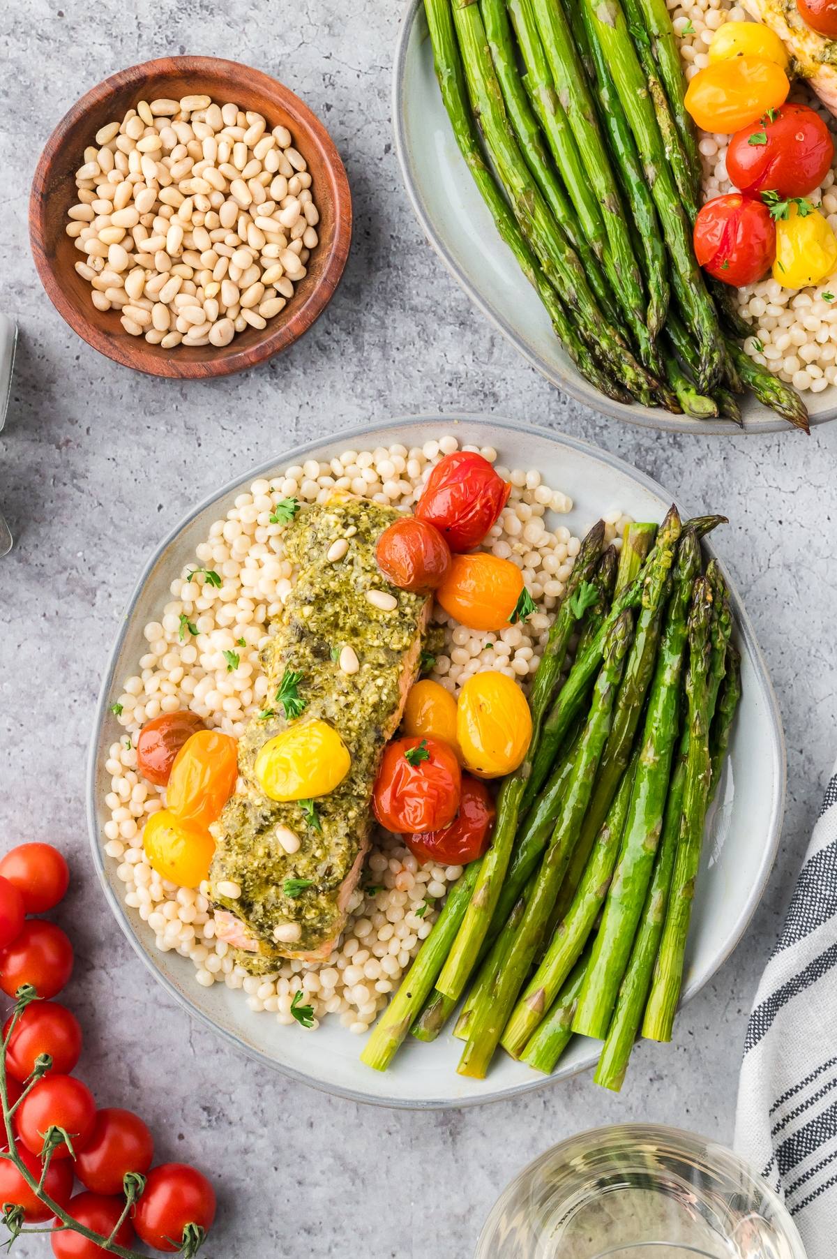 Plate with couscous, pesto salmon, roasted tomatoes, and asparagus; bowl of pine nuts and tomatoes nearby.