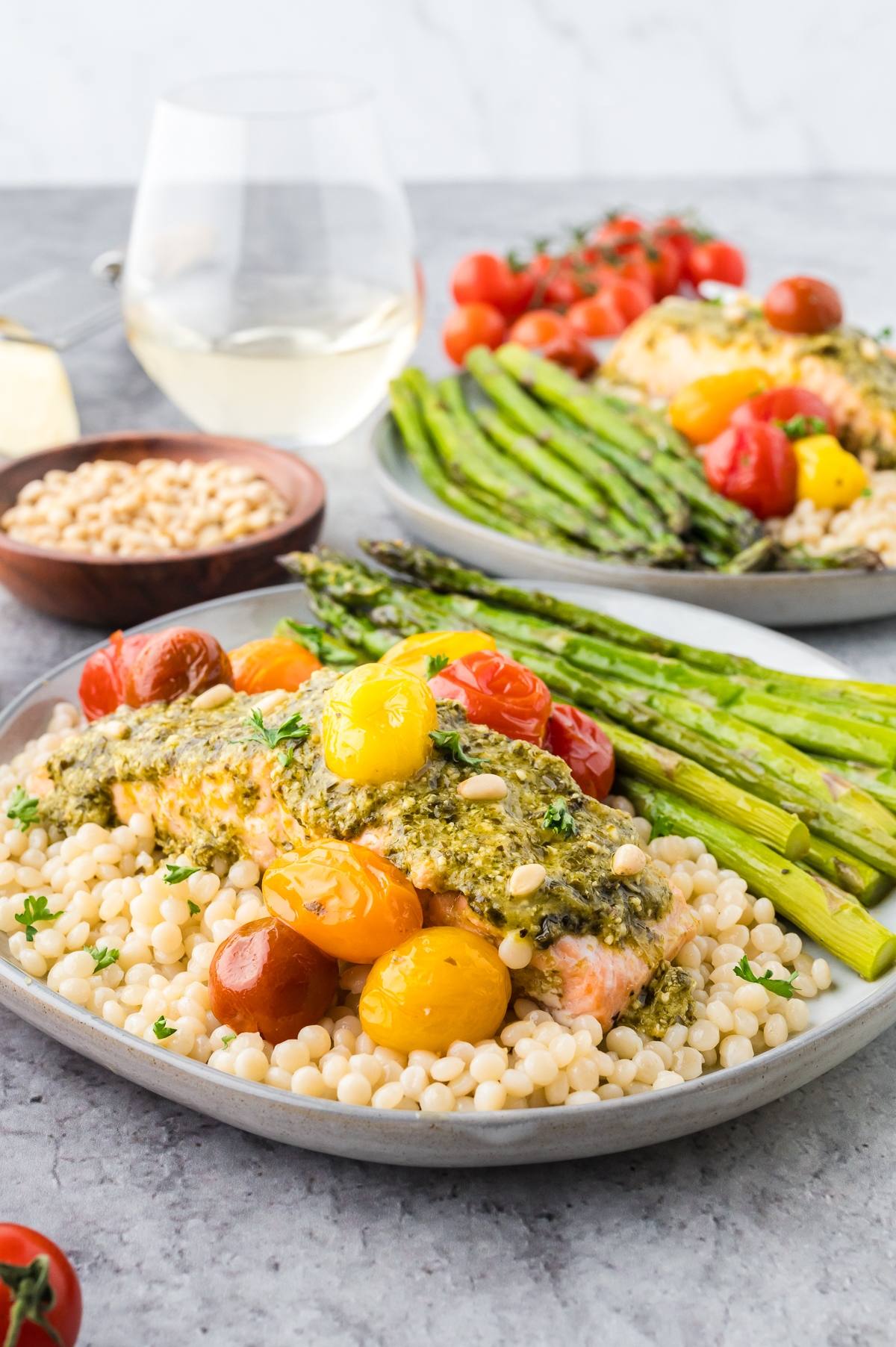 A plate with salmon, couscous, asparagus, and cherry tomatoes, with a glass of white wine in the background.