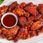 A platter of slow cooker BBQ wings glazed to perfection, served with a bowl of dipping sauce in the center and garnished with fresh herbs.