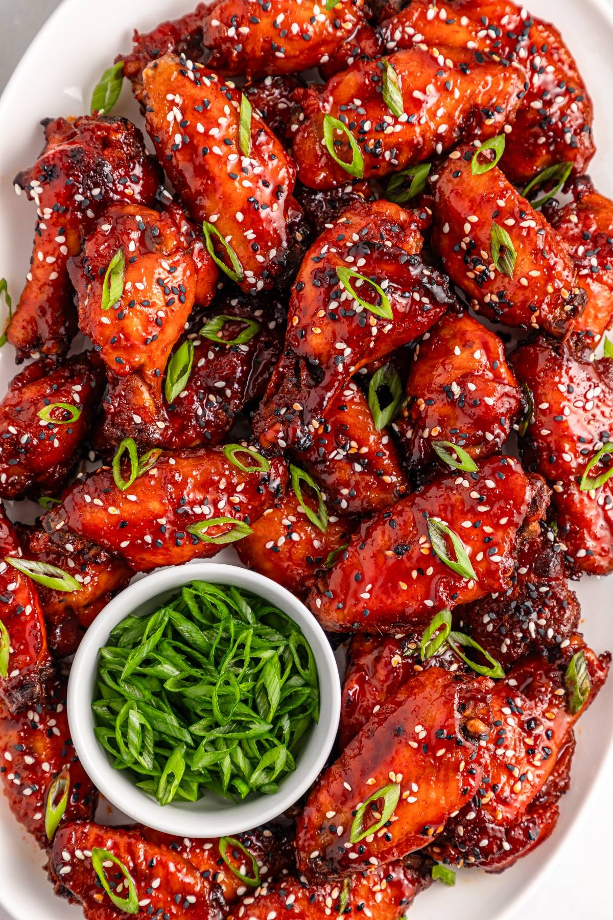 Plate of glazed chicken wings garnished with sesame seeds and sliced green onions, with a bowl of green onions.