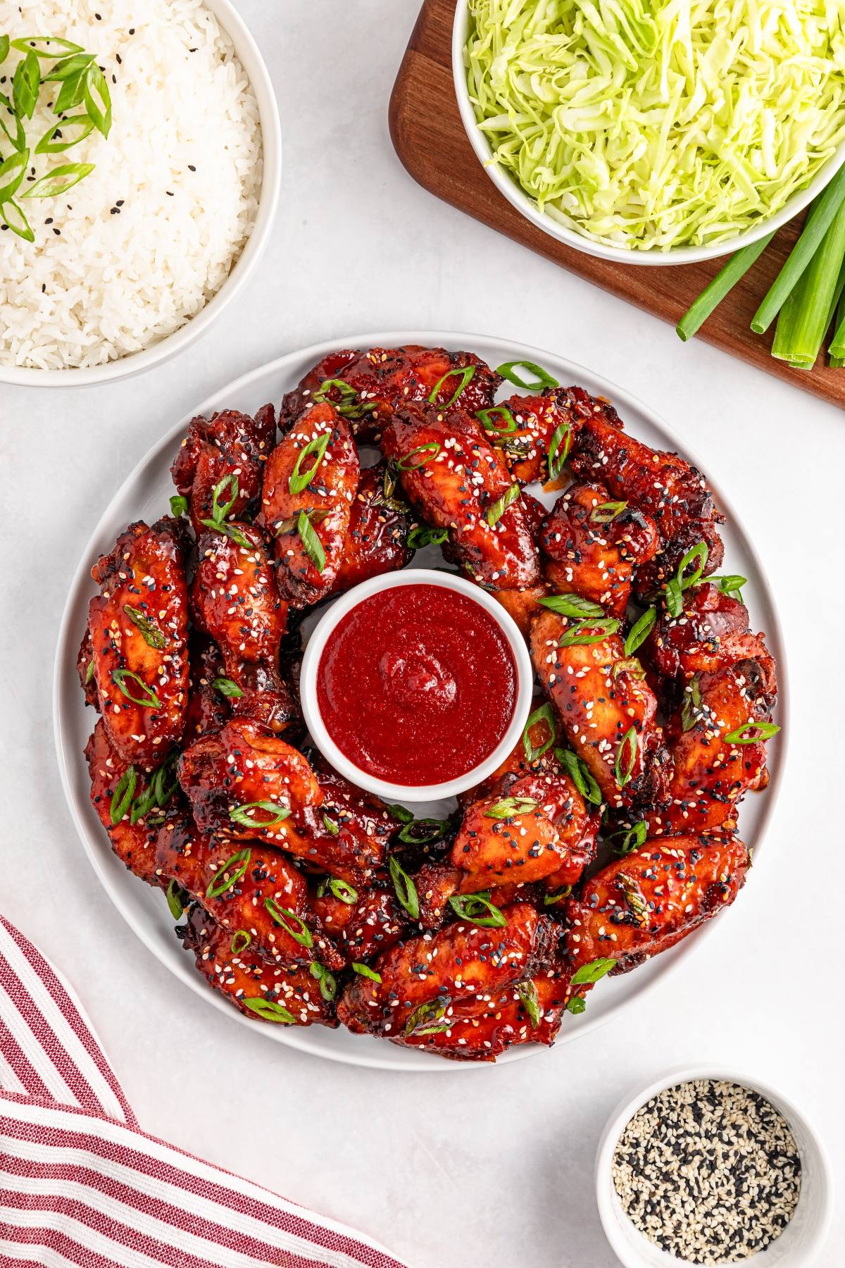 A plate of glazed chicken wings with sesame seeds and green onions, served with red dipping sauce in the center.