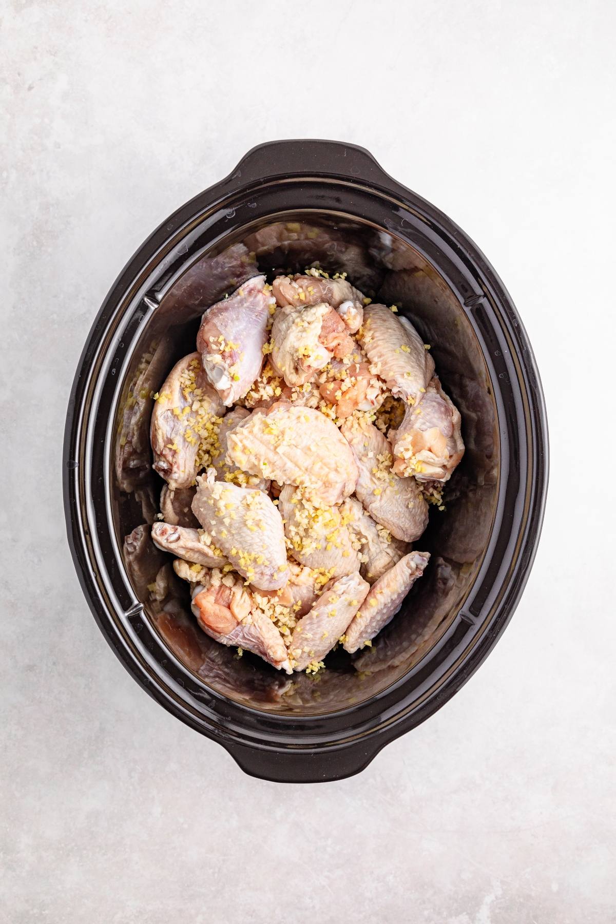 Raw chicken wings with minced garlic in a slow cooker, viewed from above on a light background.