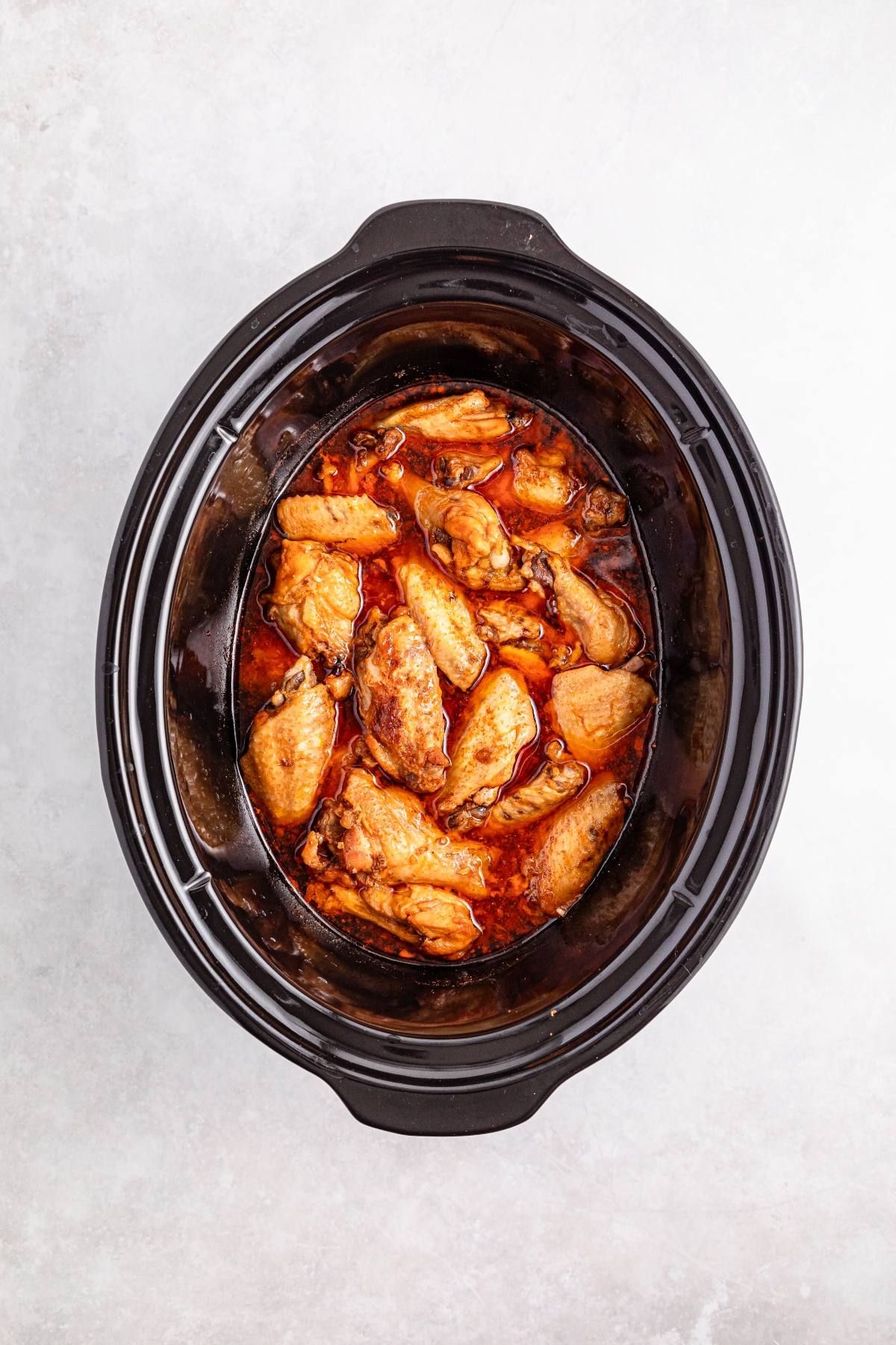 Chicken wings in a rich sauce inside a black slow cooker, viewed from above on a light surface.
