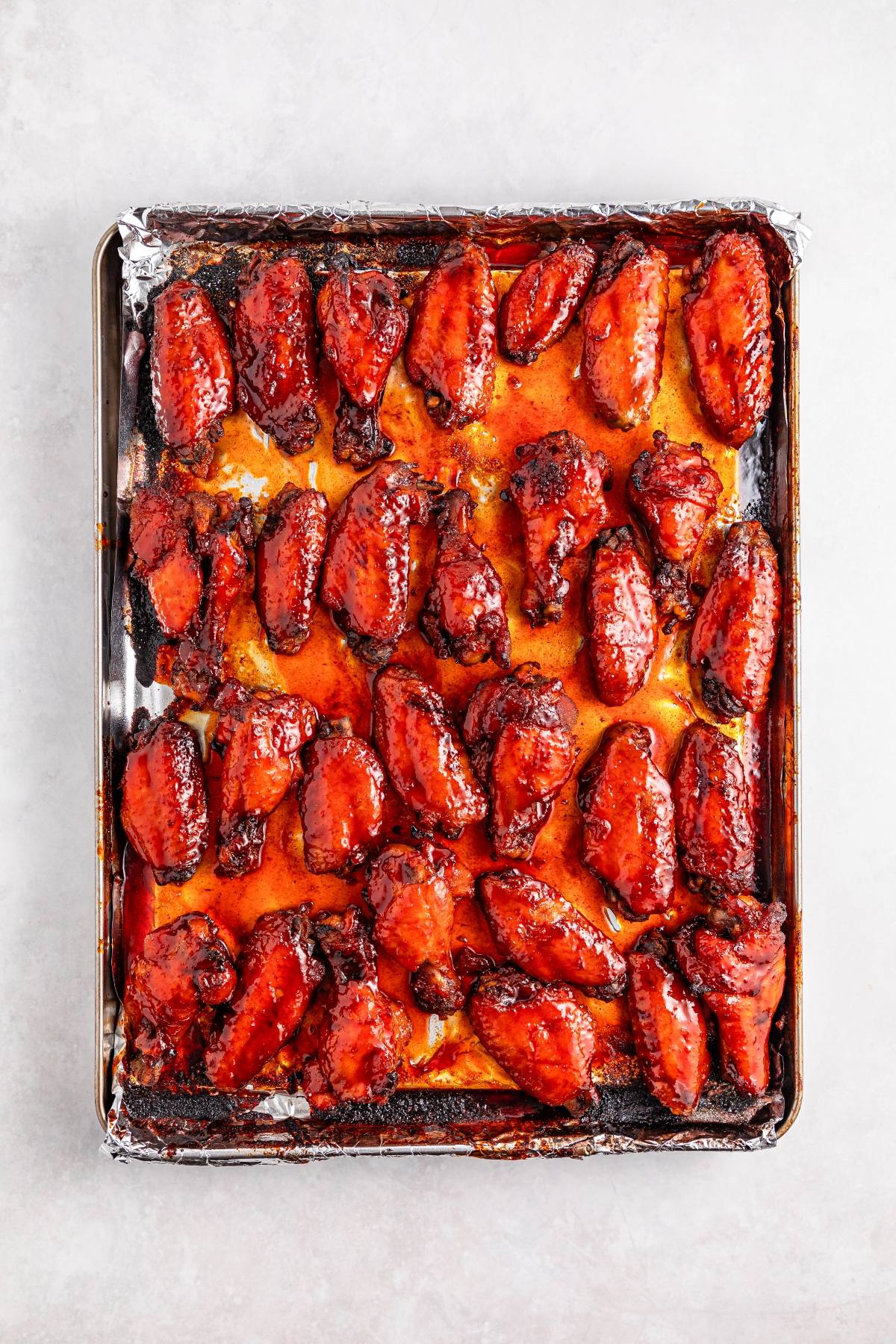 A baking sheet lined with foil, filled with glazed, baked chicken wings in a spicy red sauce.