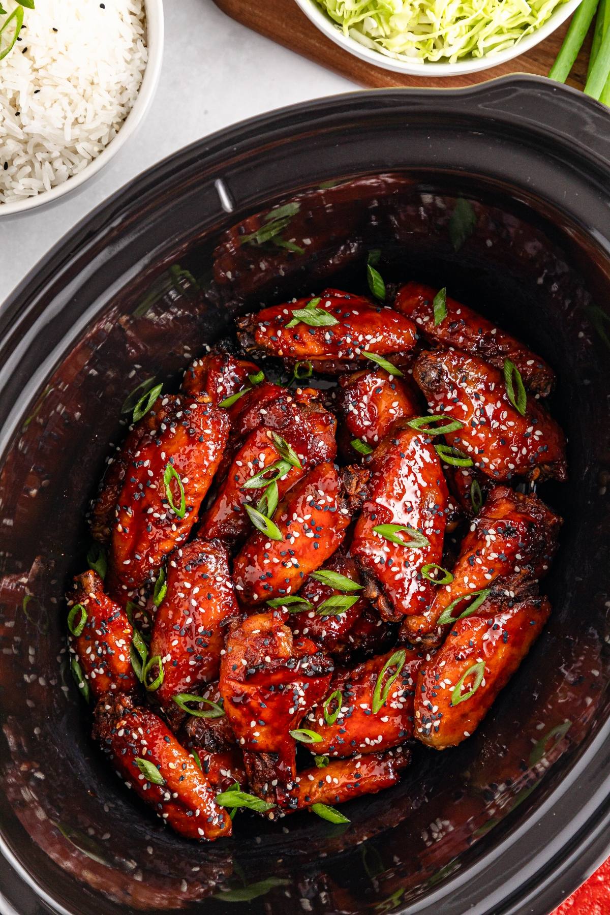 Sticky glazed chicken wings garnished with sesame seeds and green onions in a black slow cooker.