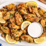 A platter of slow cooker lemon pepper wings garnished with lemon slices and parsley, served with a cup of dipping sauce.
