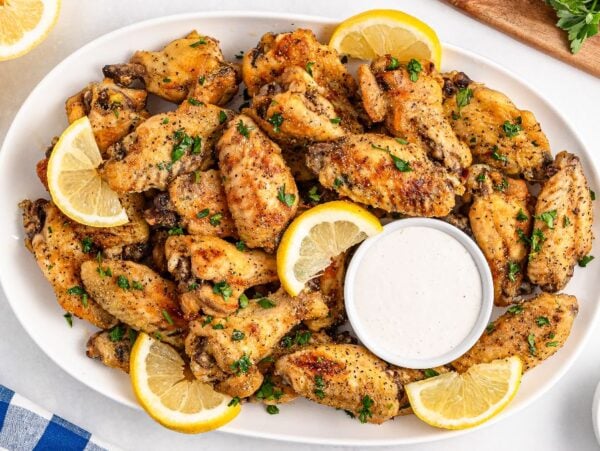 A platter of slow cooker lemon pepper wings garnished with lemon slices and parsley, served with a cup of dipping sauce.