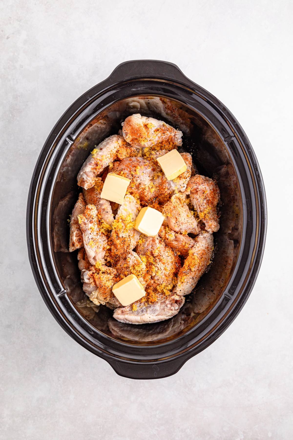 Raw seasoned chicken wings and four butter cubes in a black slow cooker on a light surface.