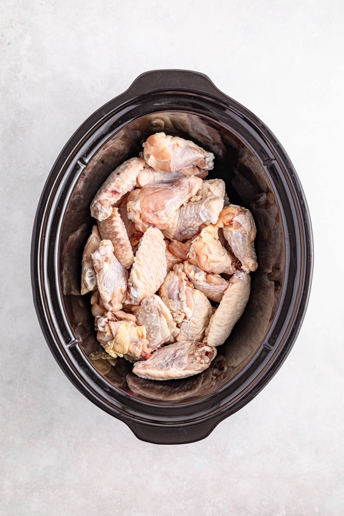 Raw chicken wings placed inside a black slow cooker, ready to be cooked.