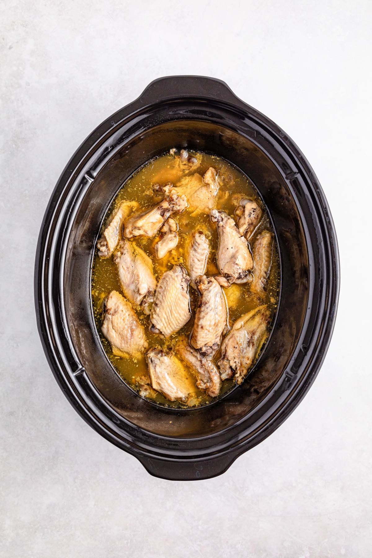 Chicken wings cooking in a slow cooker with broth, viewed from above on a light background.