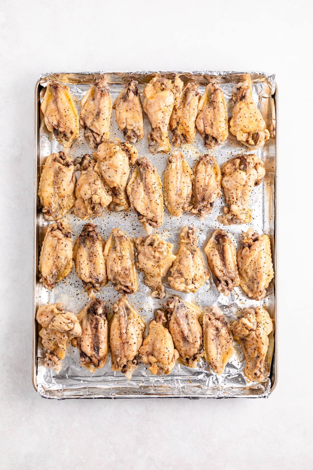 A baking sheet lined with foil holds rows of seasoned, uncooked chicken wings arranged neatly.