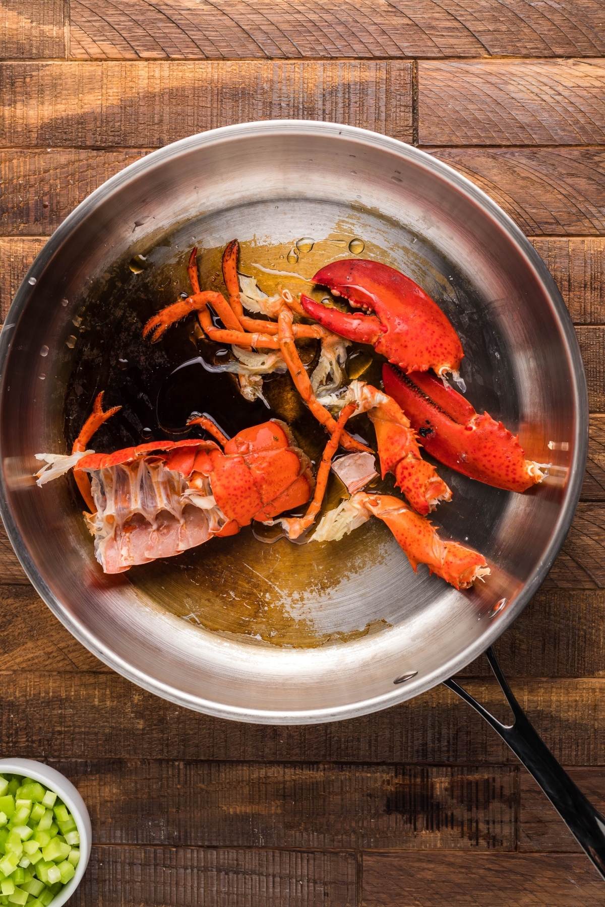 Lobster shells and claws simmer in a stainless steel pan on a wooden surface, next to a bowl of chopped celery—perfect ingredients for the best bisque recipe or a rich, creamy seafood soup.