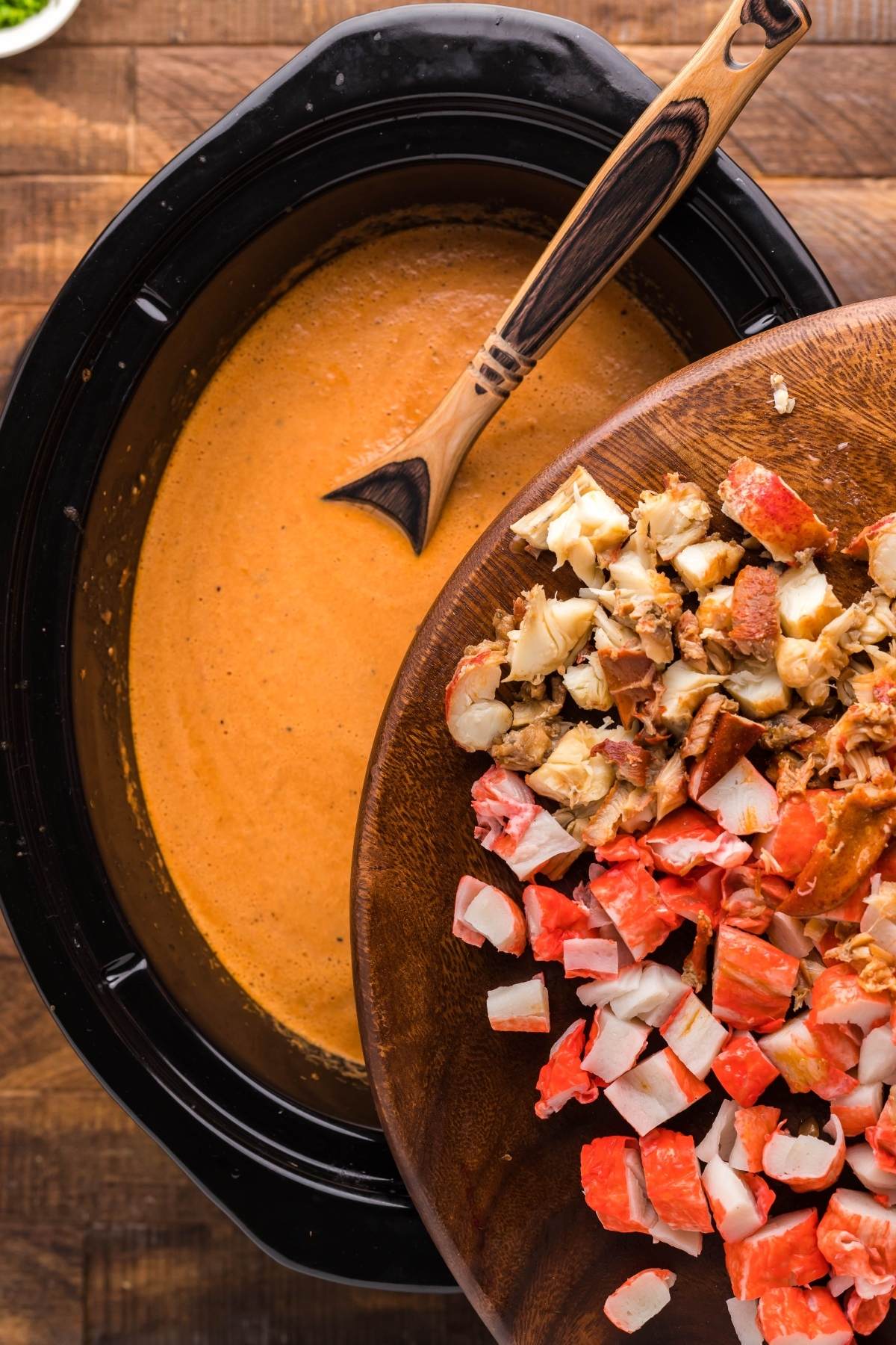 Chopped seafood being added to a creamy orange Crockpot seafood bisque with a wooden spoon.