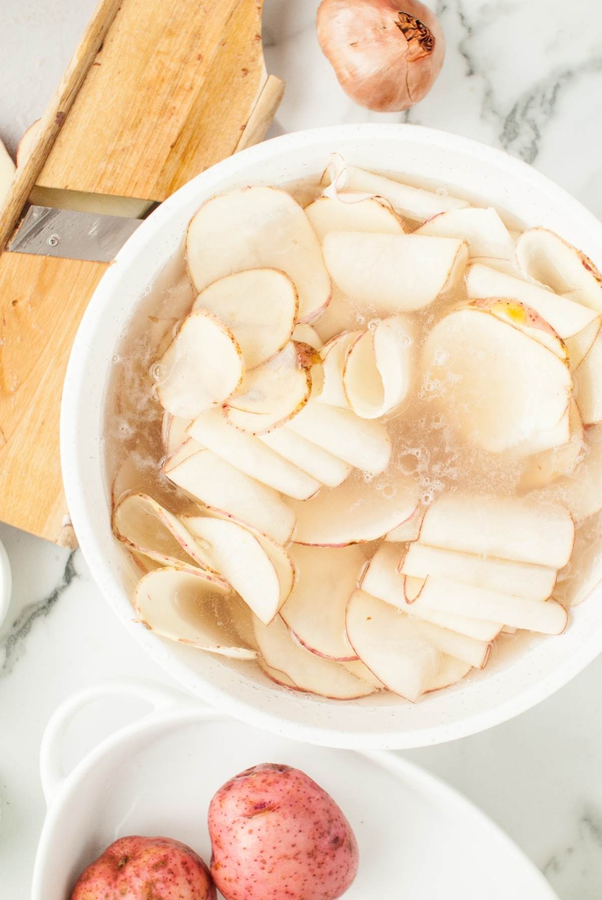 Thinly sliced potatoes soak in a bowl of water, ready for creating scalloped potatoes from scratch, with whole potatoes and a slicer nearby.