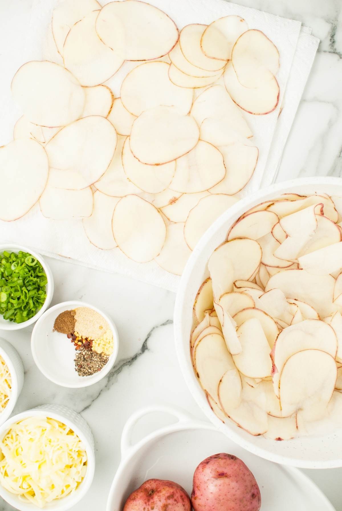 Thinly sliced red potatoes rest on a towel and in a bowl, with small bowls of cheese, spices, and green onions nearby—perfect for making gourmet scalloped potatoes from scratch.