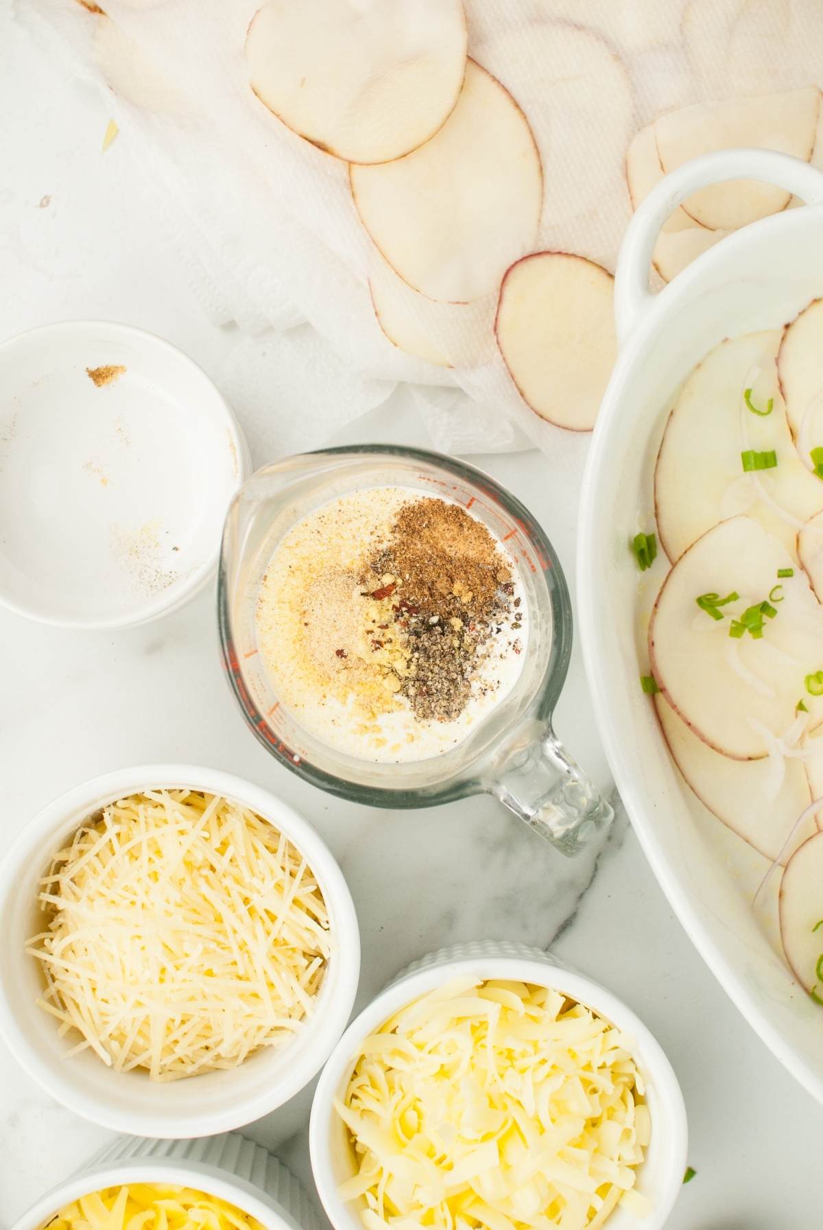 Measuring cup with spices, shredded cheese in bowls, and sliced potatoes on a white surface—perfect ingredients for making gourmet scalloped potatoes from scratch.