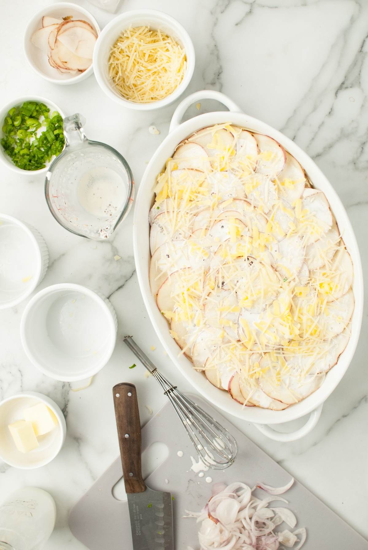 Sliced potatoes with cheese in a white baking dish, surrounded by bowls of ingredients on a marble counter—perfect for making gourmet scalloped potatoes from scratch.