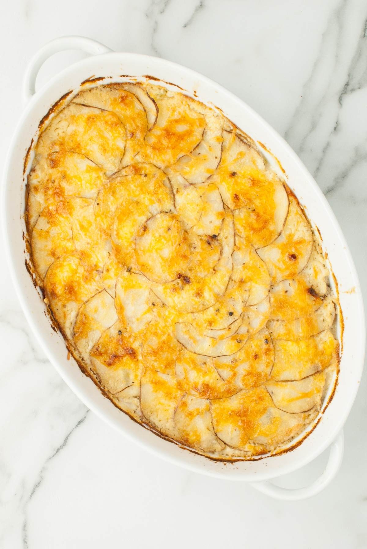 Oval dish filled with cheesy baked scalloped potatoes from scratch, topped with a golden, bubbly cheese crust for the best Potatoes Au Gratin experience.
