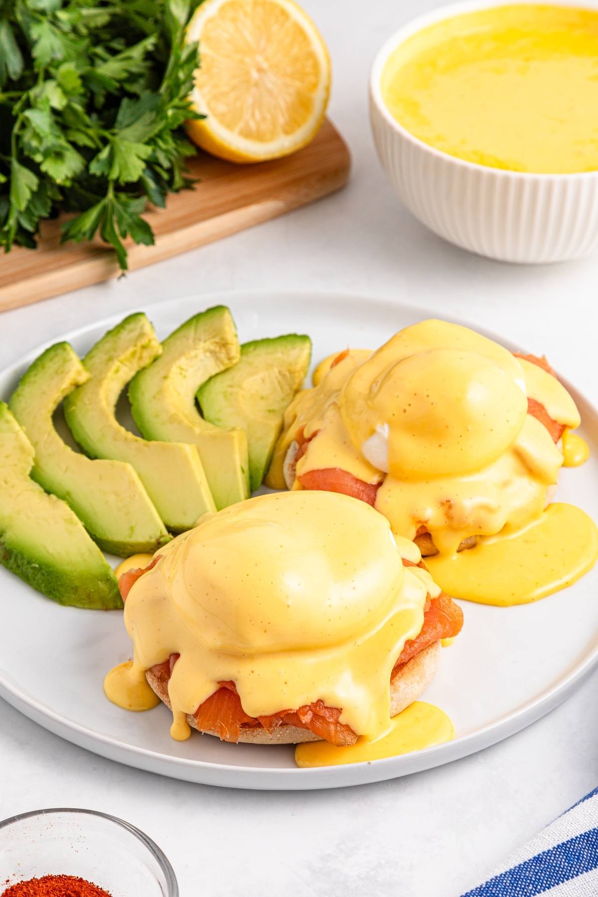 Two eggs Benedict with hollandaise sauce, sliced avocado, and greenery on a white plate.