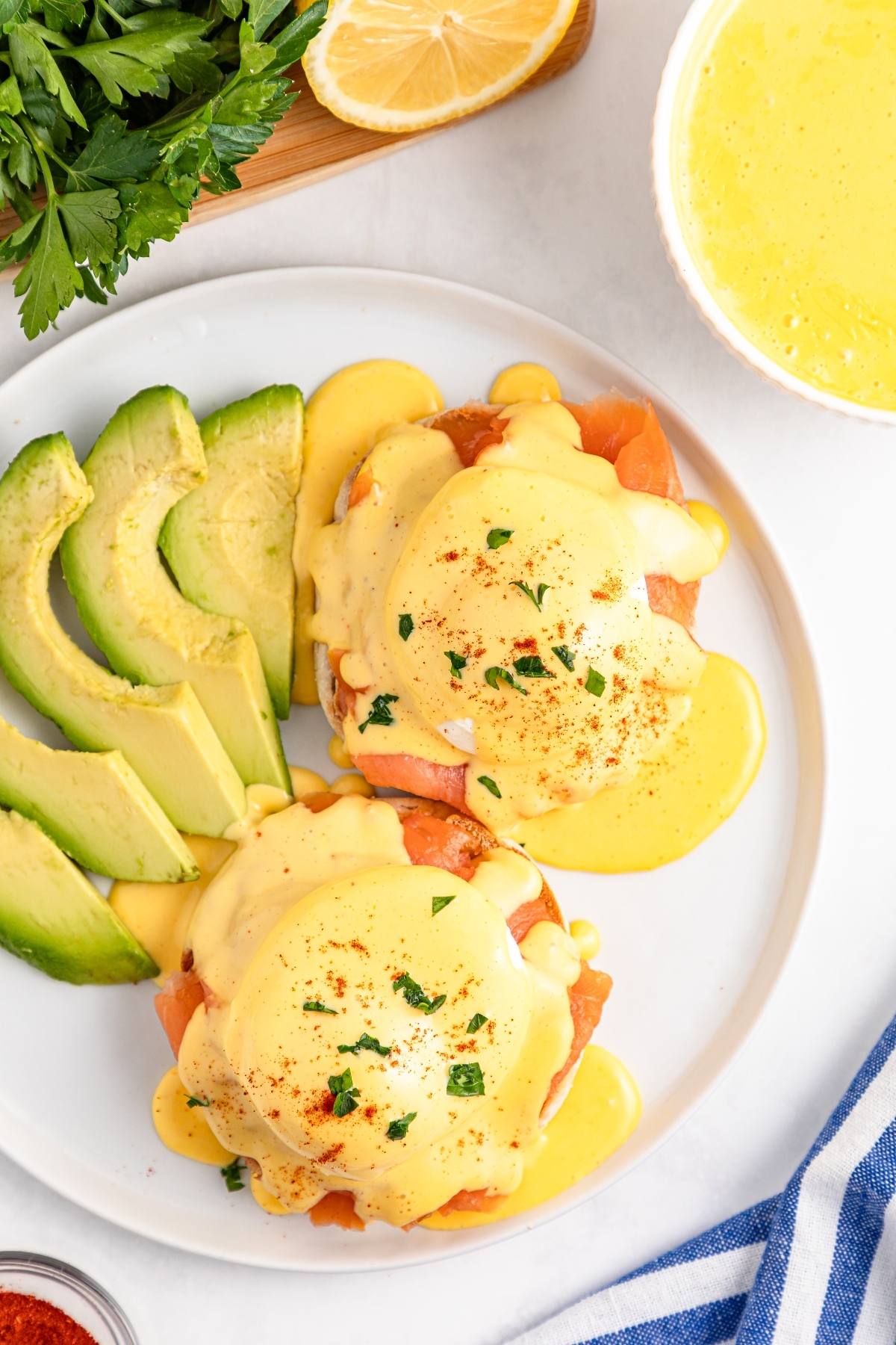 Eggs Benedict with hollandaise sauce, smoked salmon, and avocado slices on a white plate, garnished with herbs.