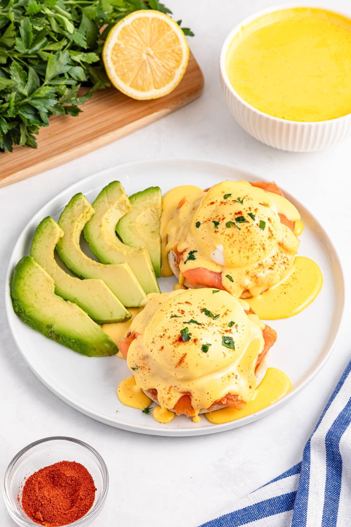Eggs Benedict with hollandaise sauce, avocado slices, herbs, and paprika on a white plate.