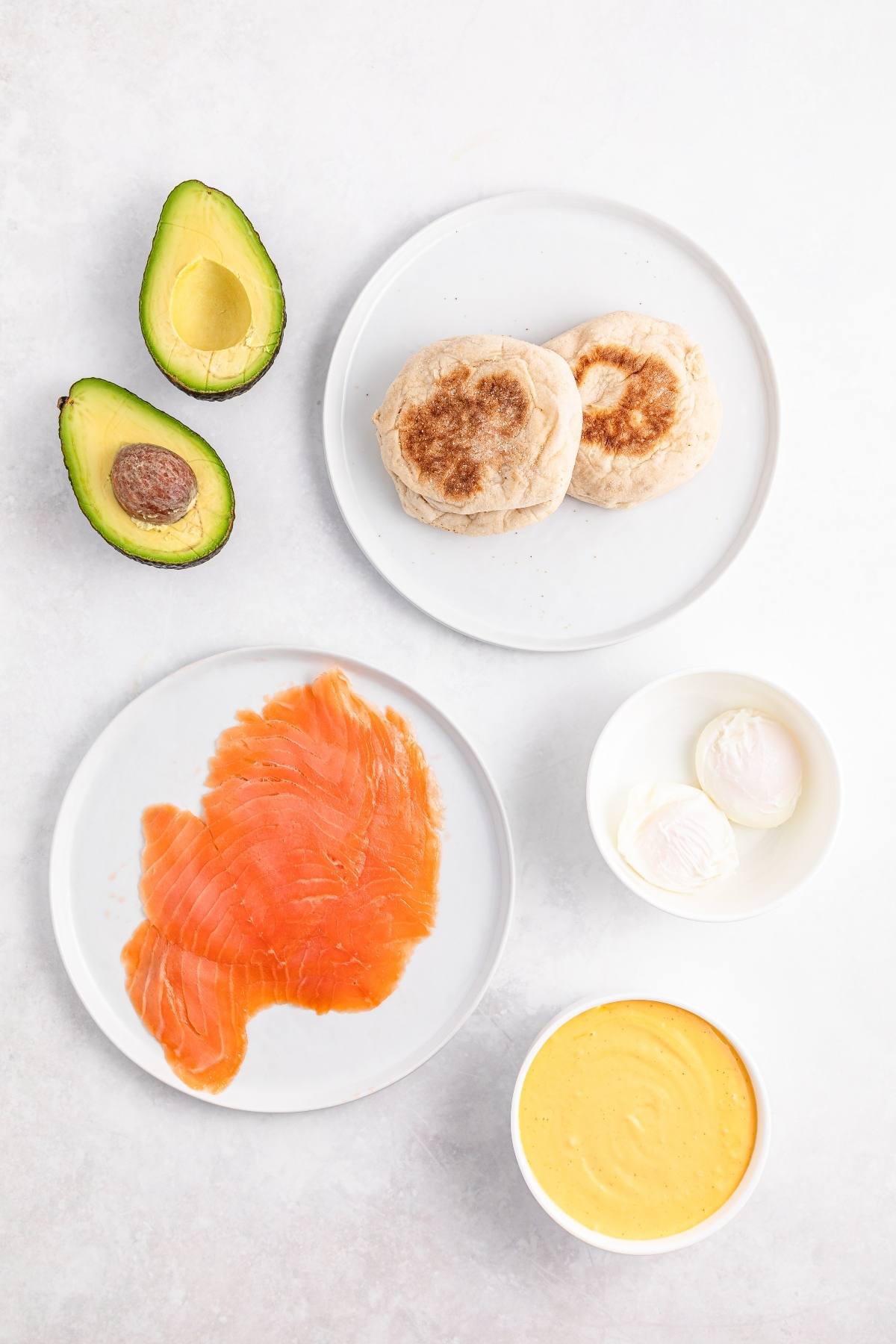 Sliced avocado, English muffins, smoked salmon, poached eggs, and hollandaise sauce on white plates.