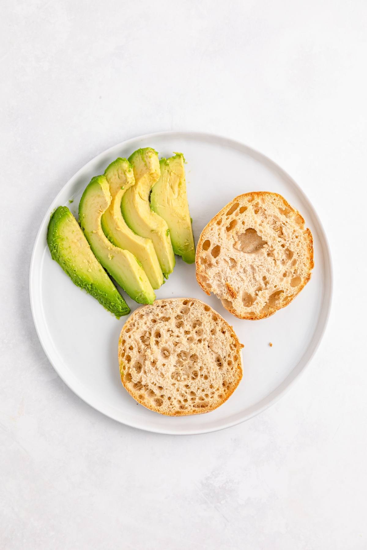 Sliced avocado and a halved bread roll on a white plate against a light background.