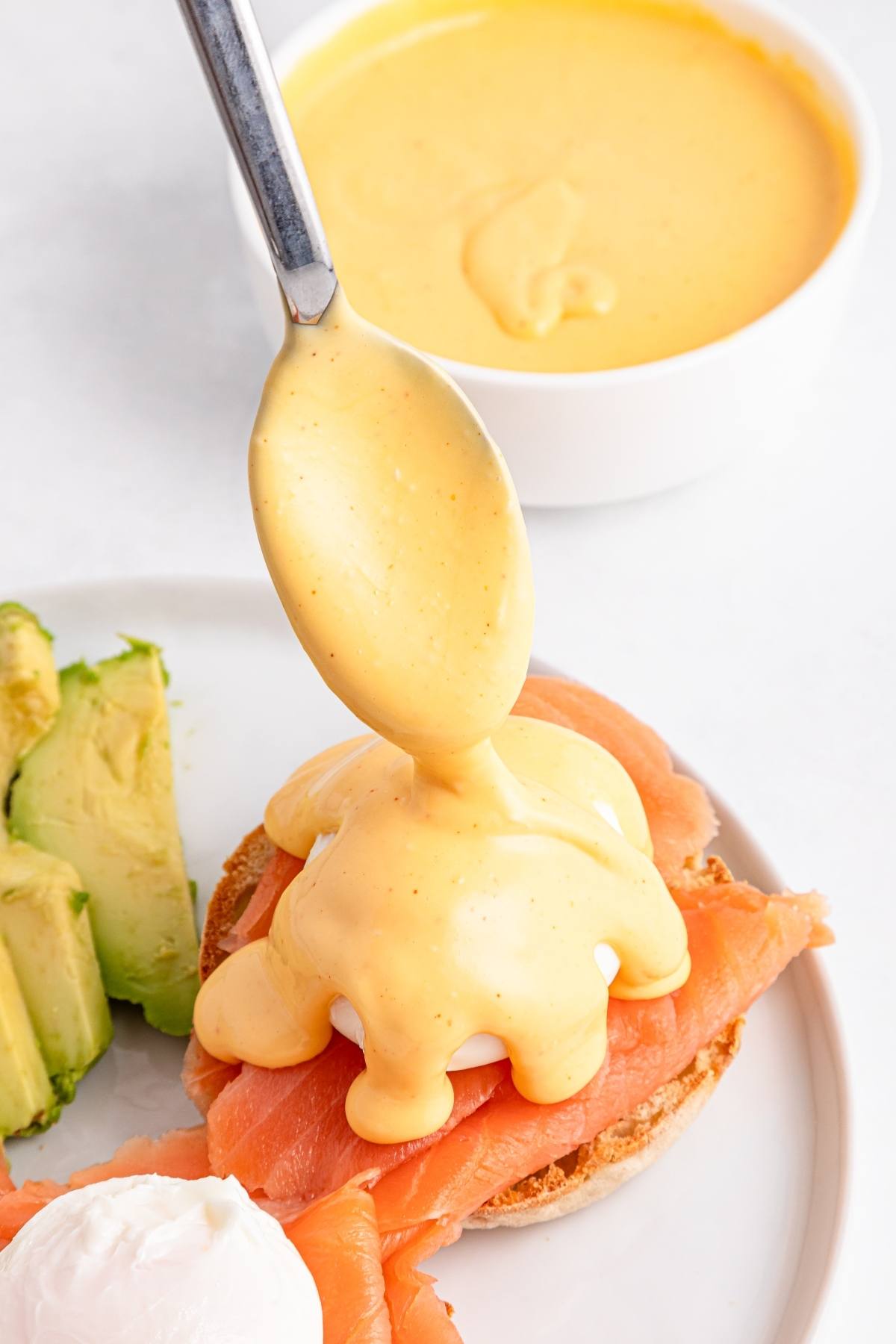 A spoon drizzling hollandaise sauce over poached eggs and smoked salmon on an English muffin, with avocado on the side.