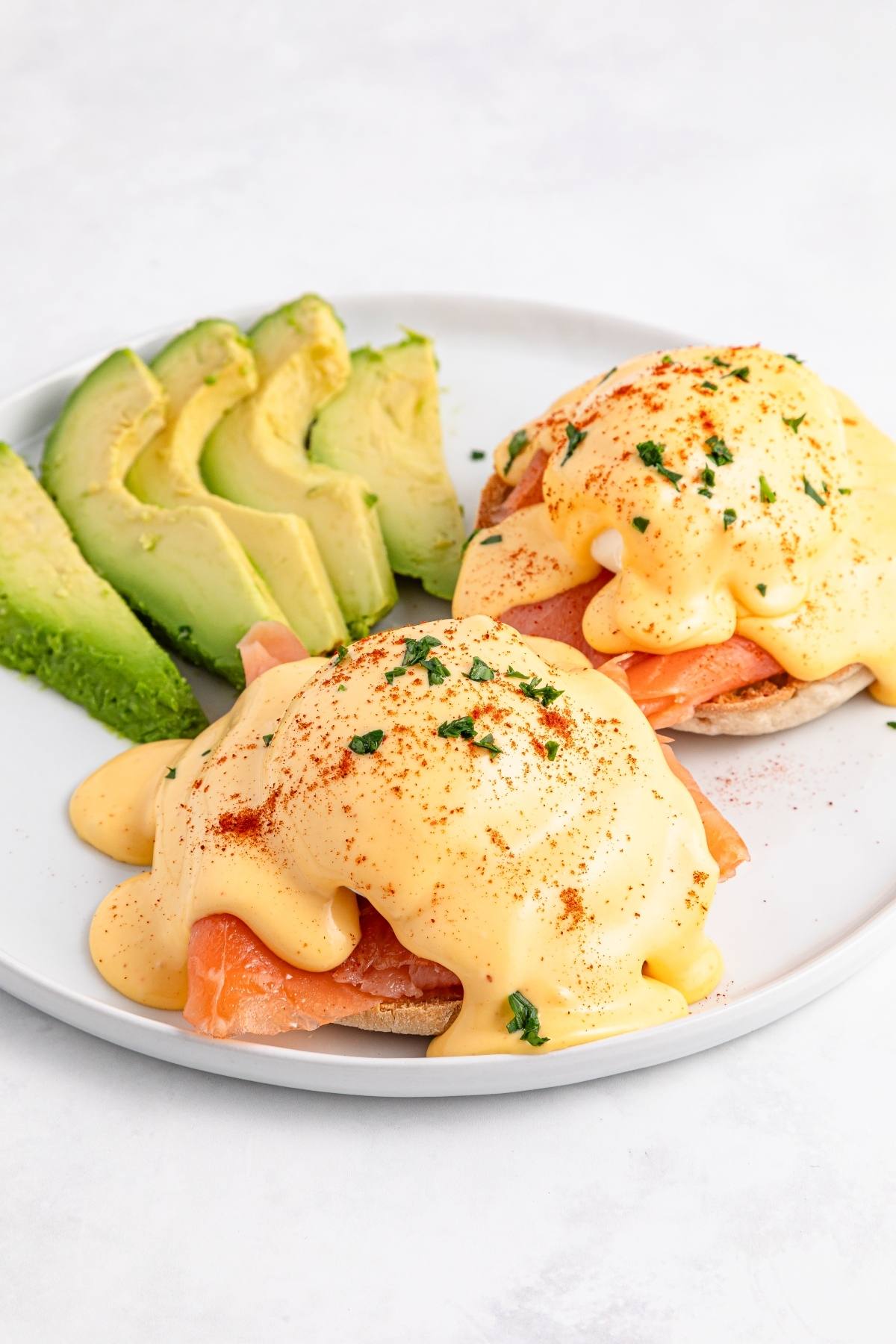 Eggs Benedict with smoked salmon and hollandaise sauce, served with sliced avocado on a white plate.