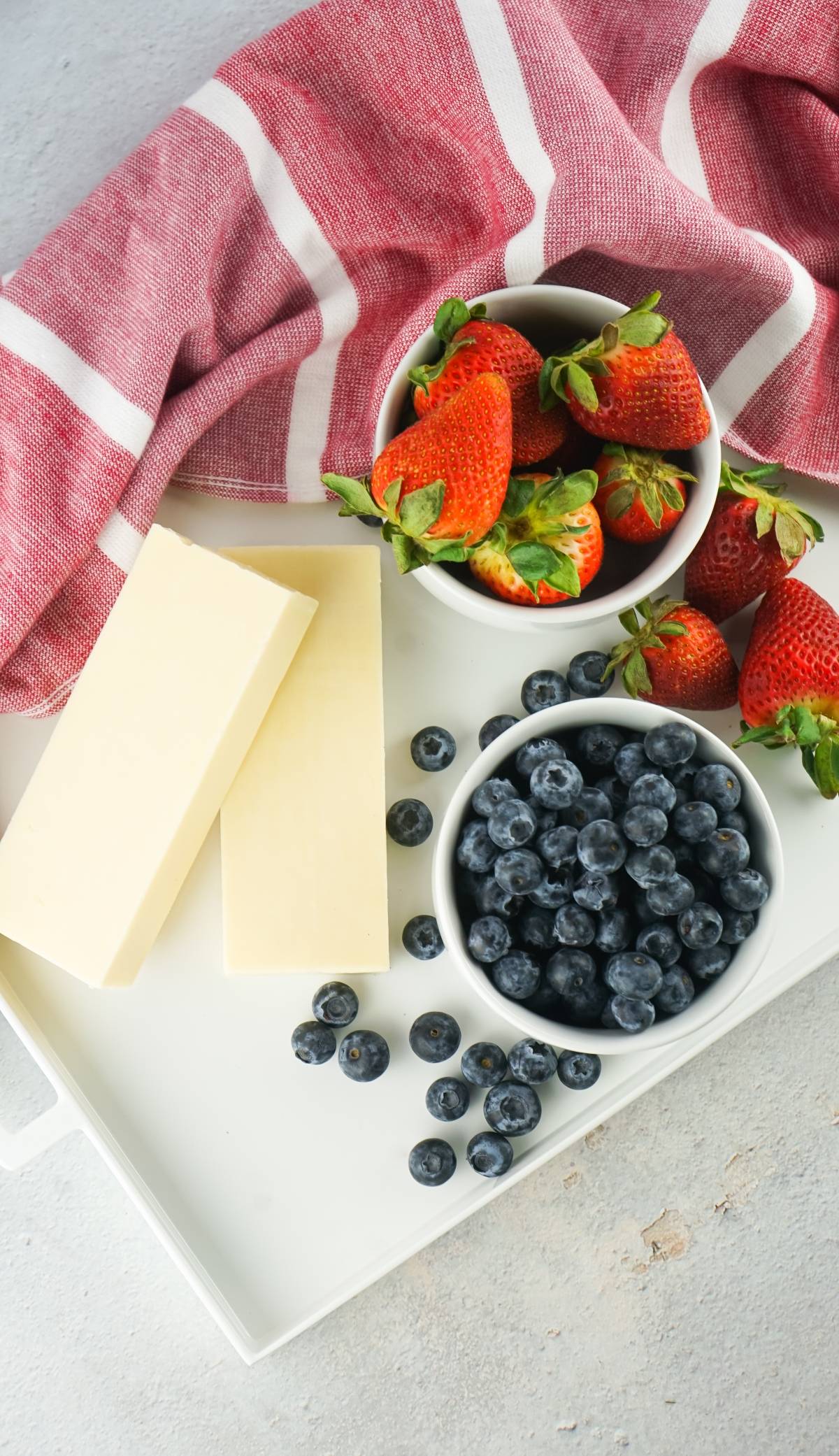Blocks of cheese, bowls of strawberries and blueberries on a white tray with a red striped cloth.