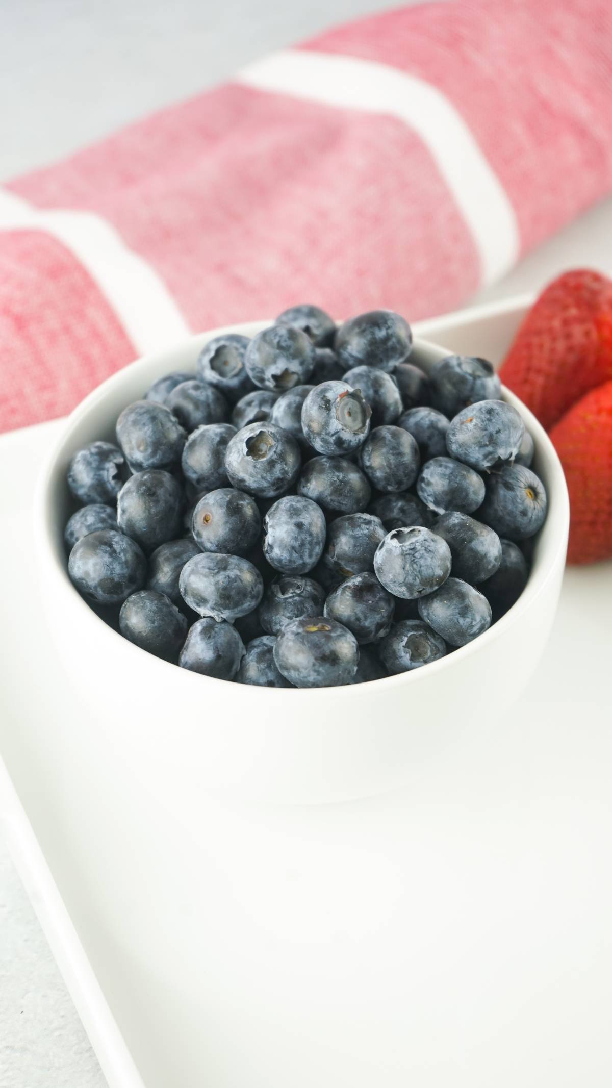 A white bowl filled with blueberries next to strawberries on a white tray with a red-striped cloth in the background.