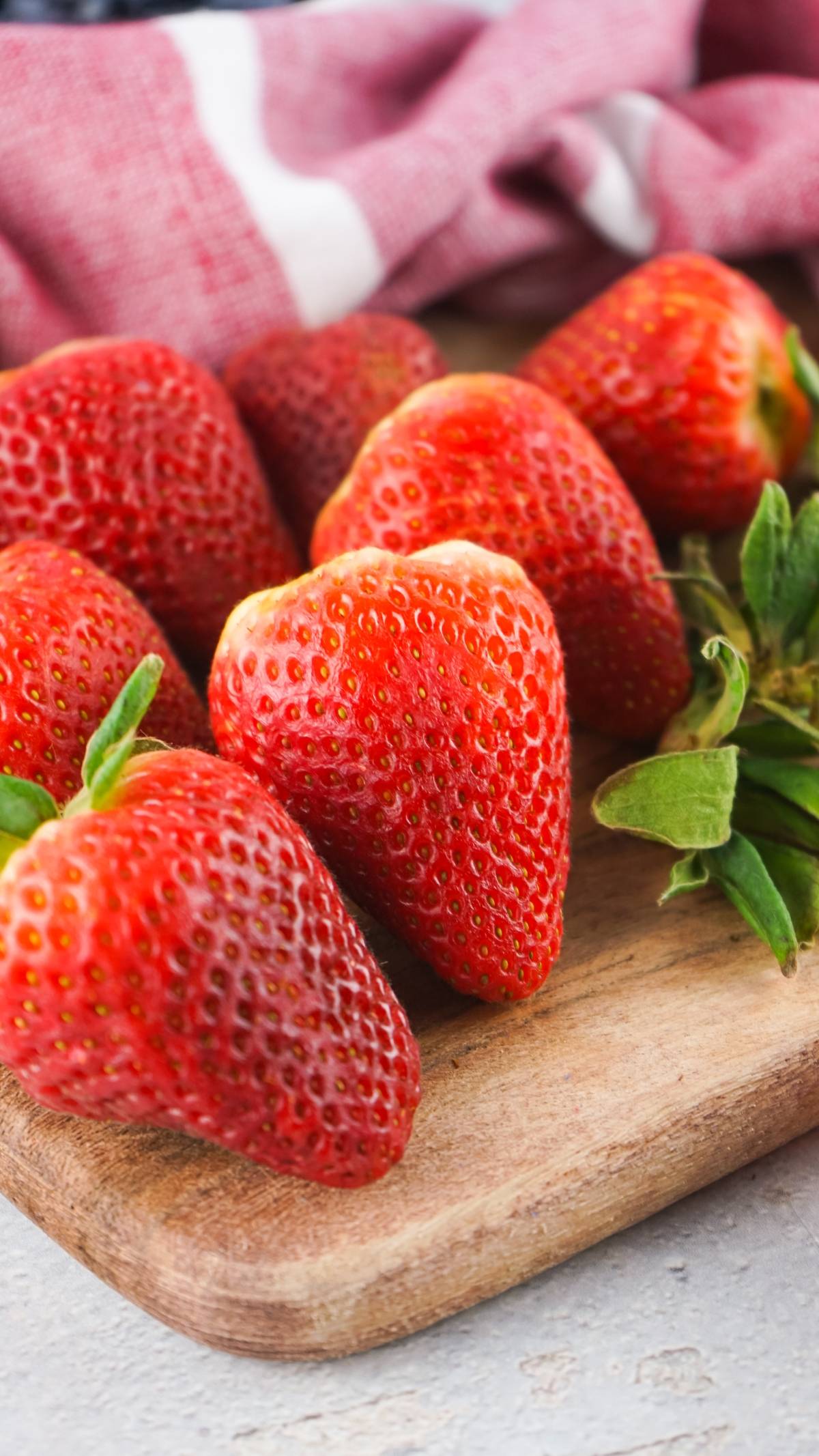 Fresh strawberries with green leaves on a wooden board, with a red and white cloth in the background.