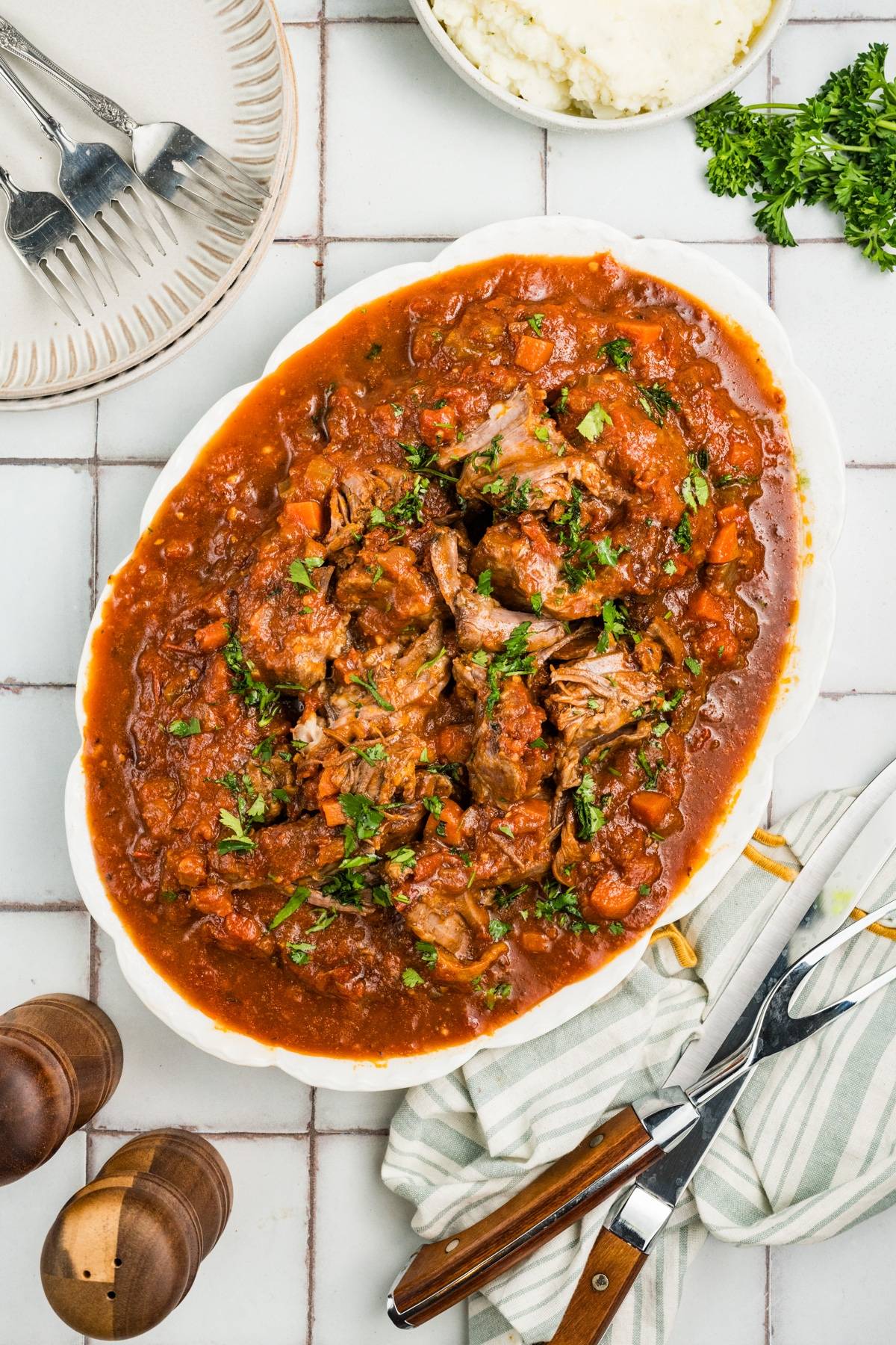 A white plate with beef stew in tomato sauce, garnished with herbs, on a tiled table with utensils nearby.