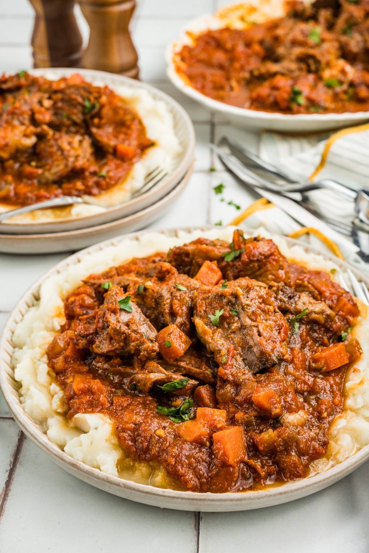 A plate of mashed potatoes topped with beef stew in tomato sauce, with carrots and herbs.