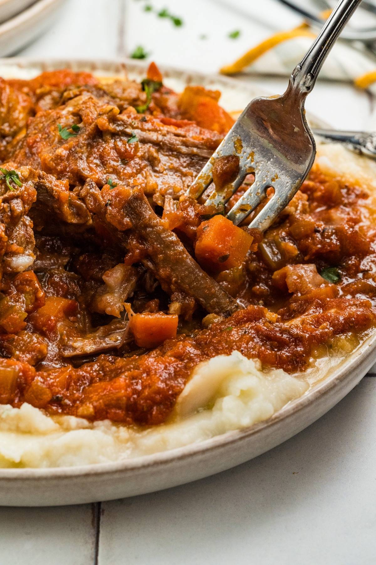 A fork lifts braised beef with vegetables and tomato sauce over creamy mashed potatoes.