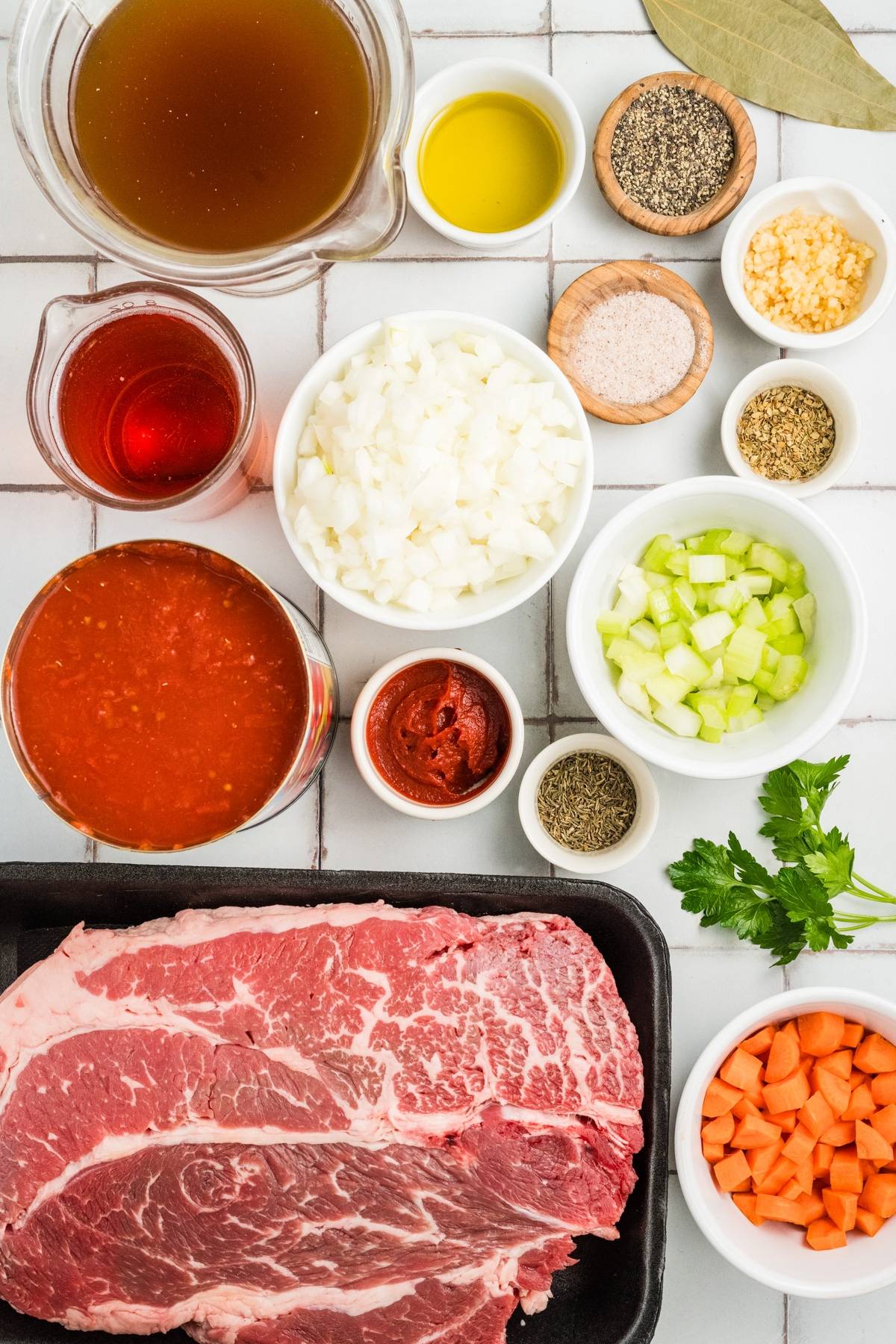 Raw beef, chopped vegetables, spices, tomato sauce, broth, and oil arranged on a white tiled surface.